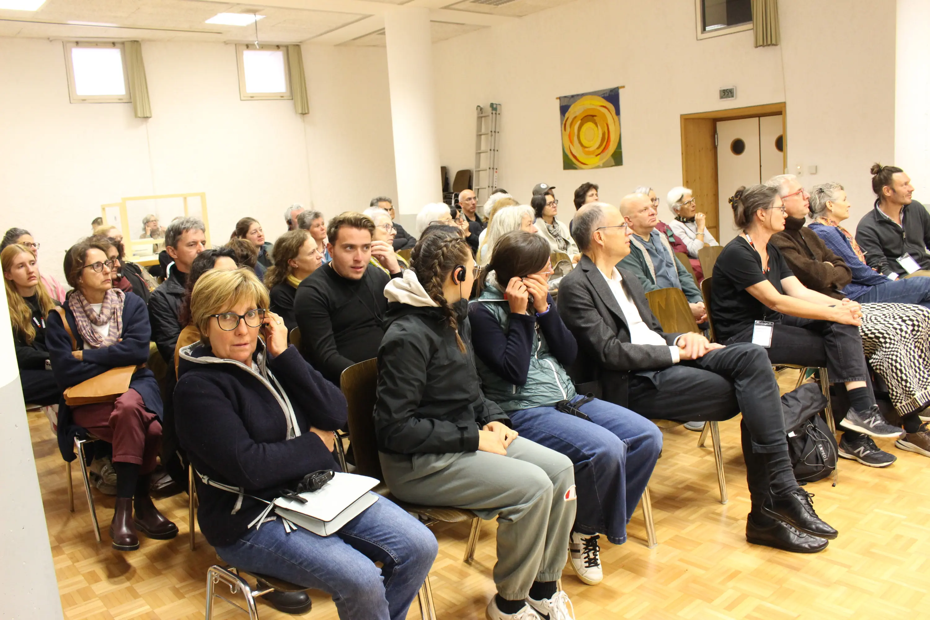 Una sala con molte persone sedute, alcune in ascolto, altre che conversano. Si nota una varietà di espressioni e stili di abbigliamento. Sullo sfondo ci sono opere d'arte appese alle pareti.