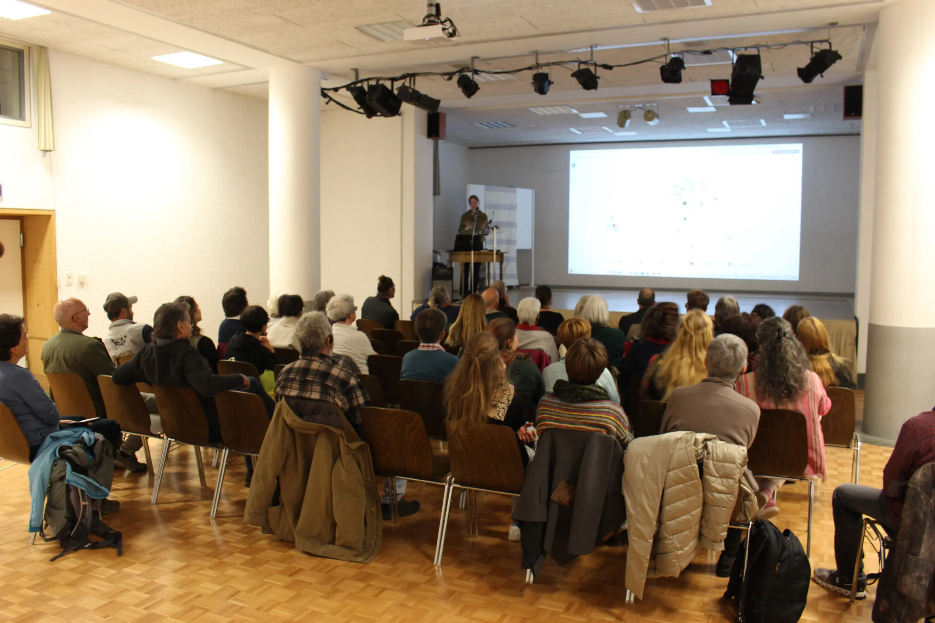 Una sala conferenze con un relatore che presenta davanti a un pubblico seduto. La stanza è illuminata e le persone ascoltano attentamente, alcune prendendo appunti.