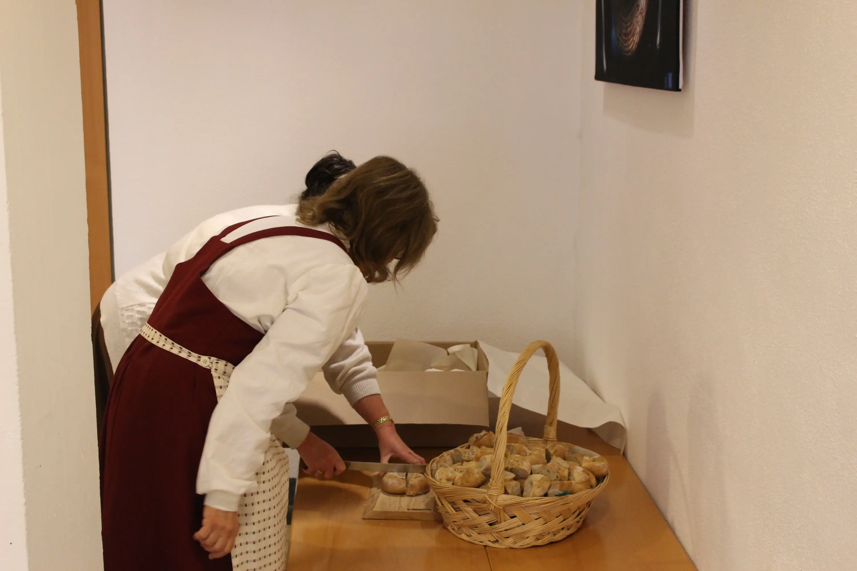 Due donne preparano del pane in un ambiente interno, una di spalle mentre affetta e l'altra si occupa di un cestino di pane su un tavolo di legno.