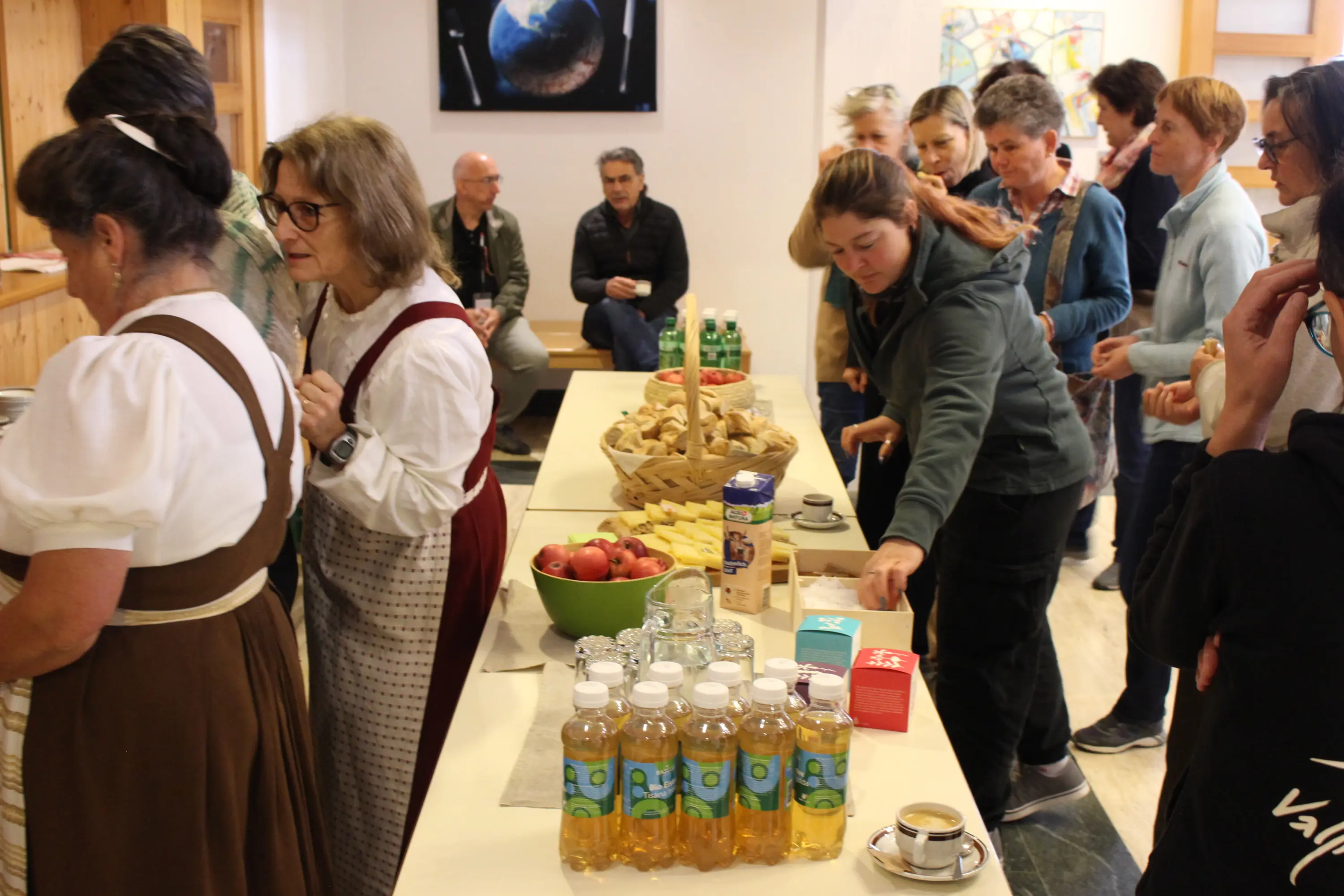 Una folla di persone in una sala, alcune vestite con abiti tradizionali. C'è un tavolo con bevande, snack e frutta. Sullo sfondo, si vedono persone che chiacchierano e un video che mostra un'immagine della Terra.