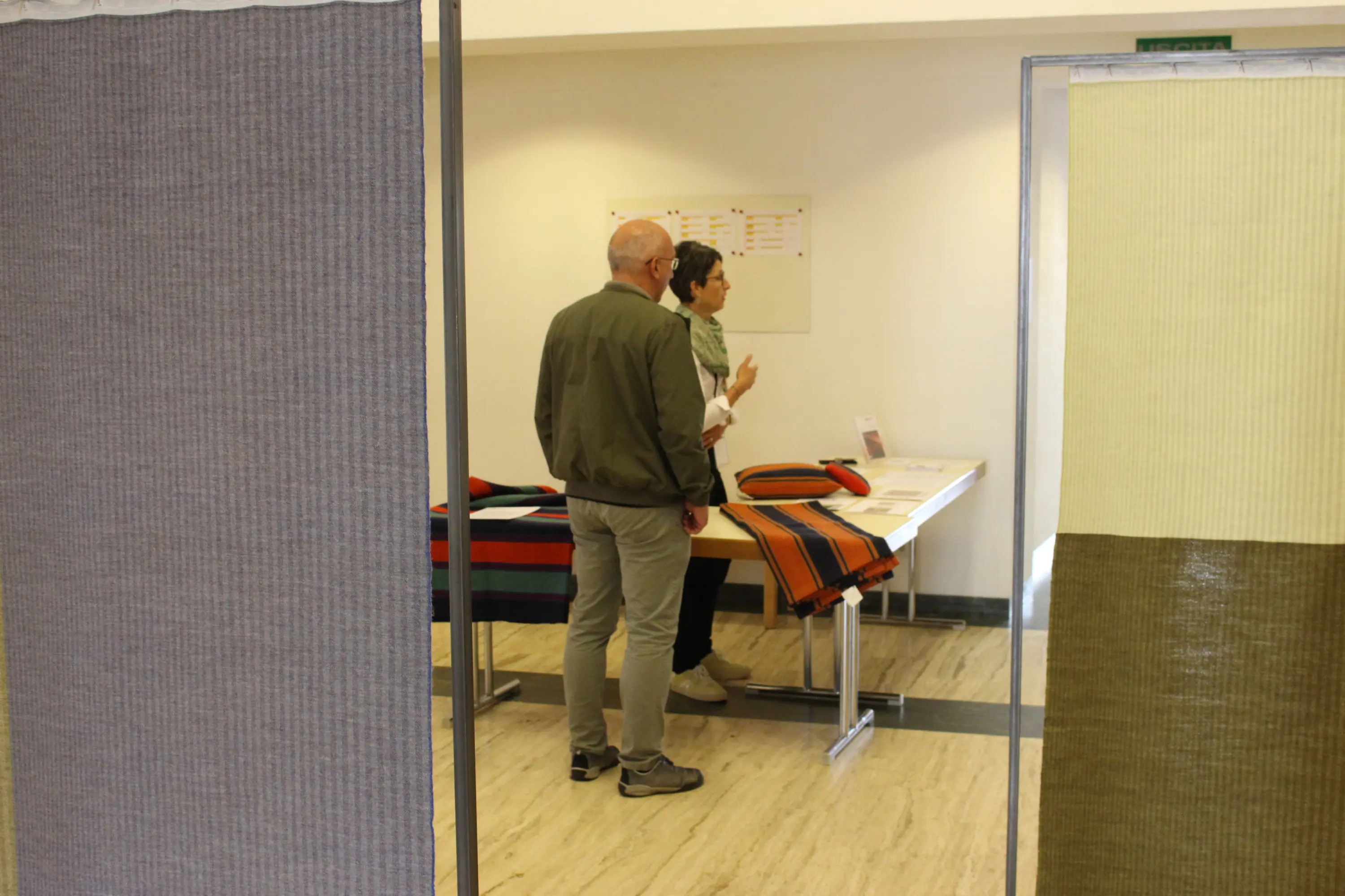 Due persone discutono davanti a un tavolo con tessuti esposti in un ambiente luminoso. I tessuti mostrano vari colori e trame.