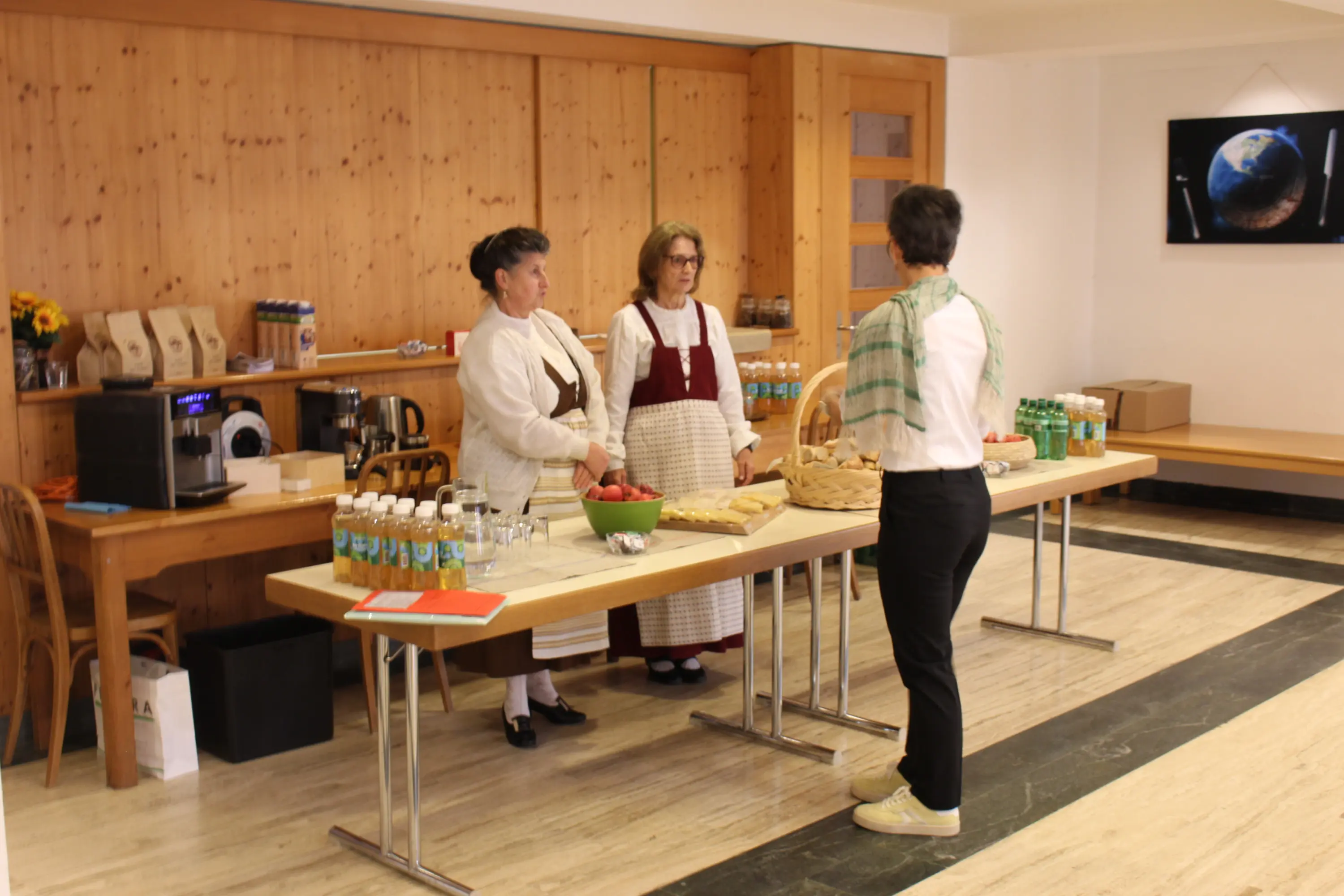 Tre donne in abiti tradizionali si trovano dietro un tavolo imbandito con cibo e bevande. Una di loro sta servendo da un grande cestino, mentre le altre due attendono. Sullo sfondo, una parete in legno e una vista di