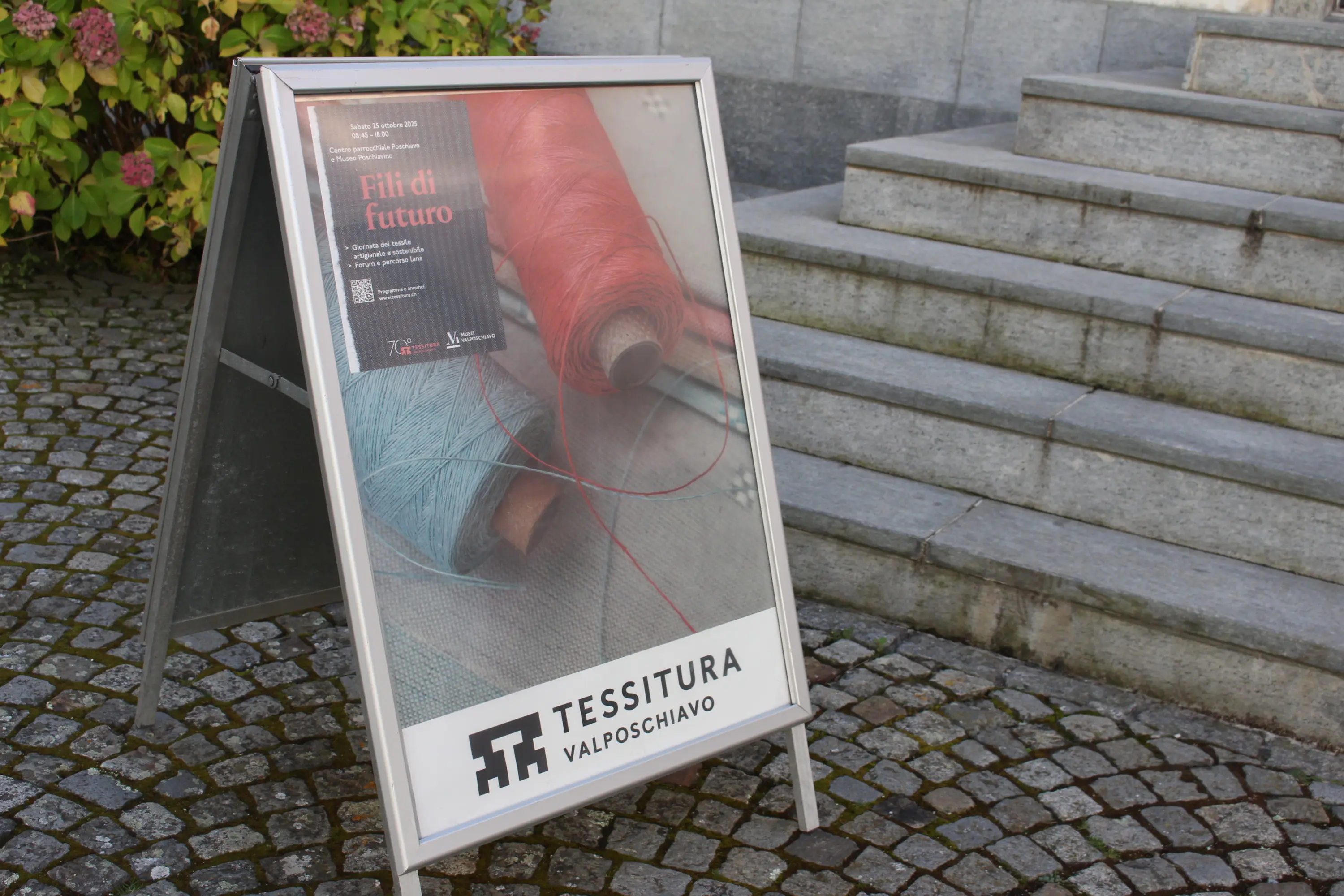 Un'insegna di legno con immagini di fili rossi e blu, promuovendo un evento intitolato "Fili di futuro". È posizionata su una superficie pavimentata accanto a dei gradini.