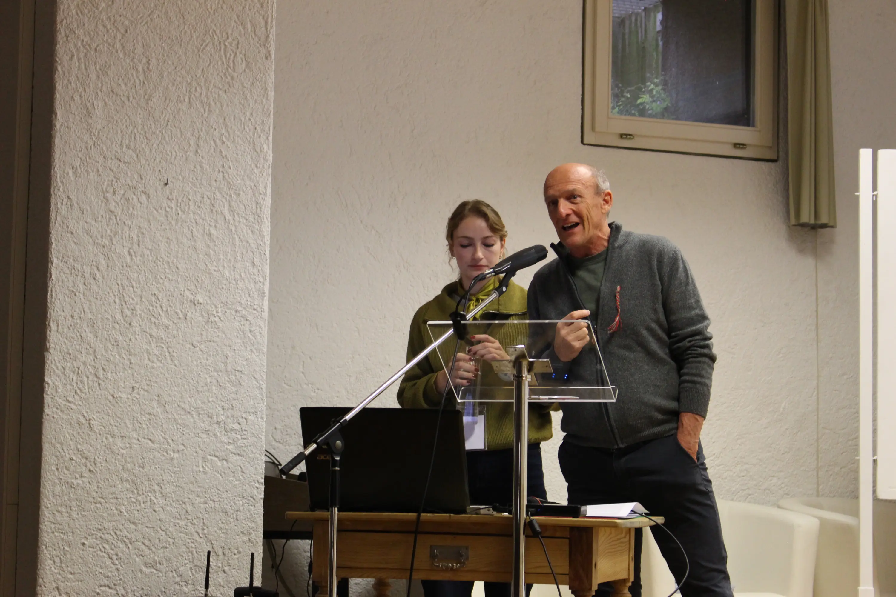 Un uomo e una giovane donna sono in piedi dietro un leggio, mentre parlano a un microfono. L'uomo indossa un maglione grigio e la donna ha un maglione verde. Dietro di loro, si vedono un computer portatile e un pro