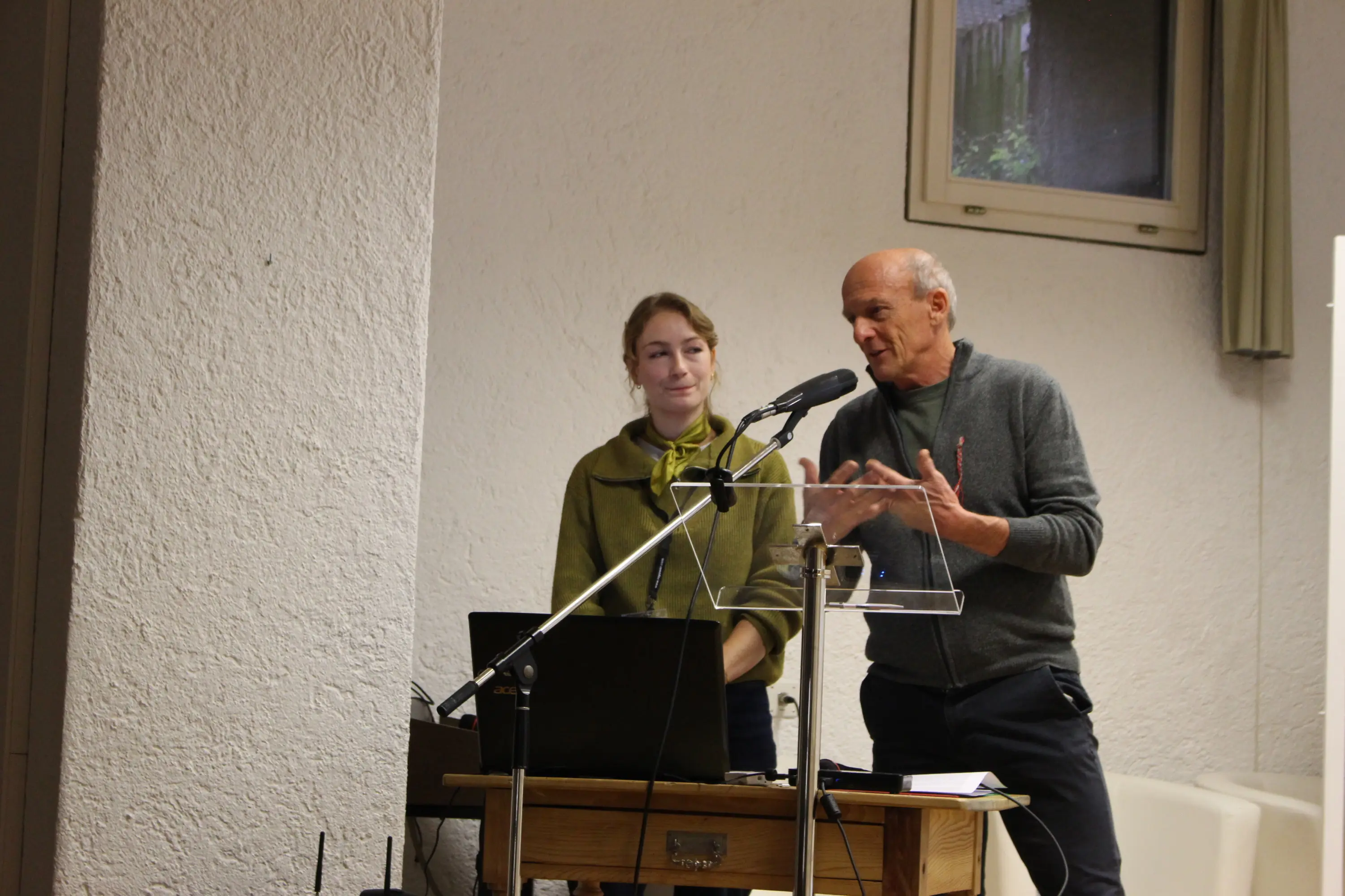 Due relatori sono in piedi dietro un leggio, uno sta parlando mentre l'altro lo ascolta attentamente. C'è un computer portatile sul tavolo davanti a loro e un microfono. L'ambiente appare luminoso e semplice.