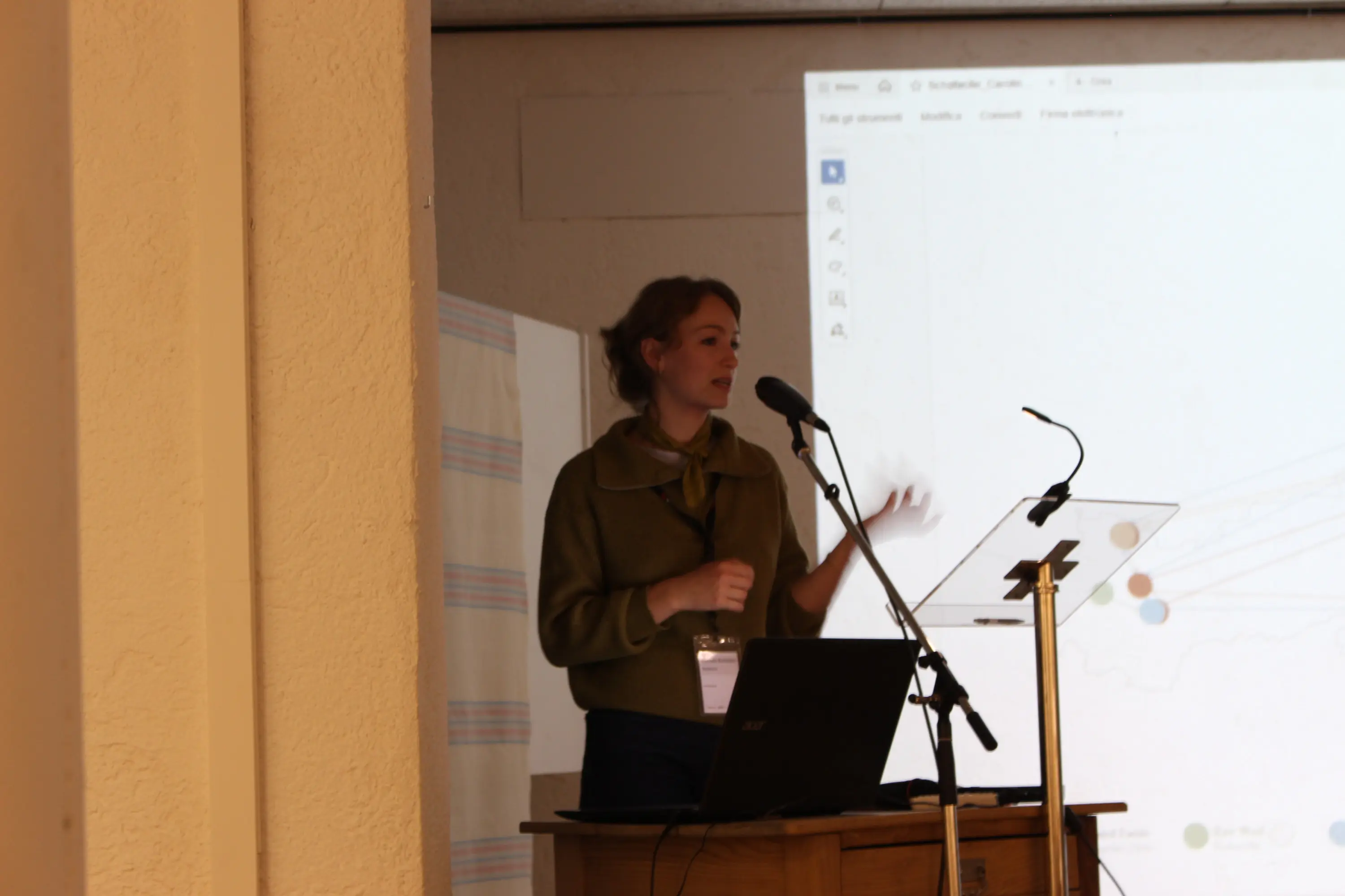 Una relatrice presenta un argomento di fronte a un pubblico, con un microfono e un supporto per appunti. Sullo sfondo è visibile una presentazione su uno schermo.