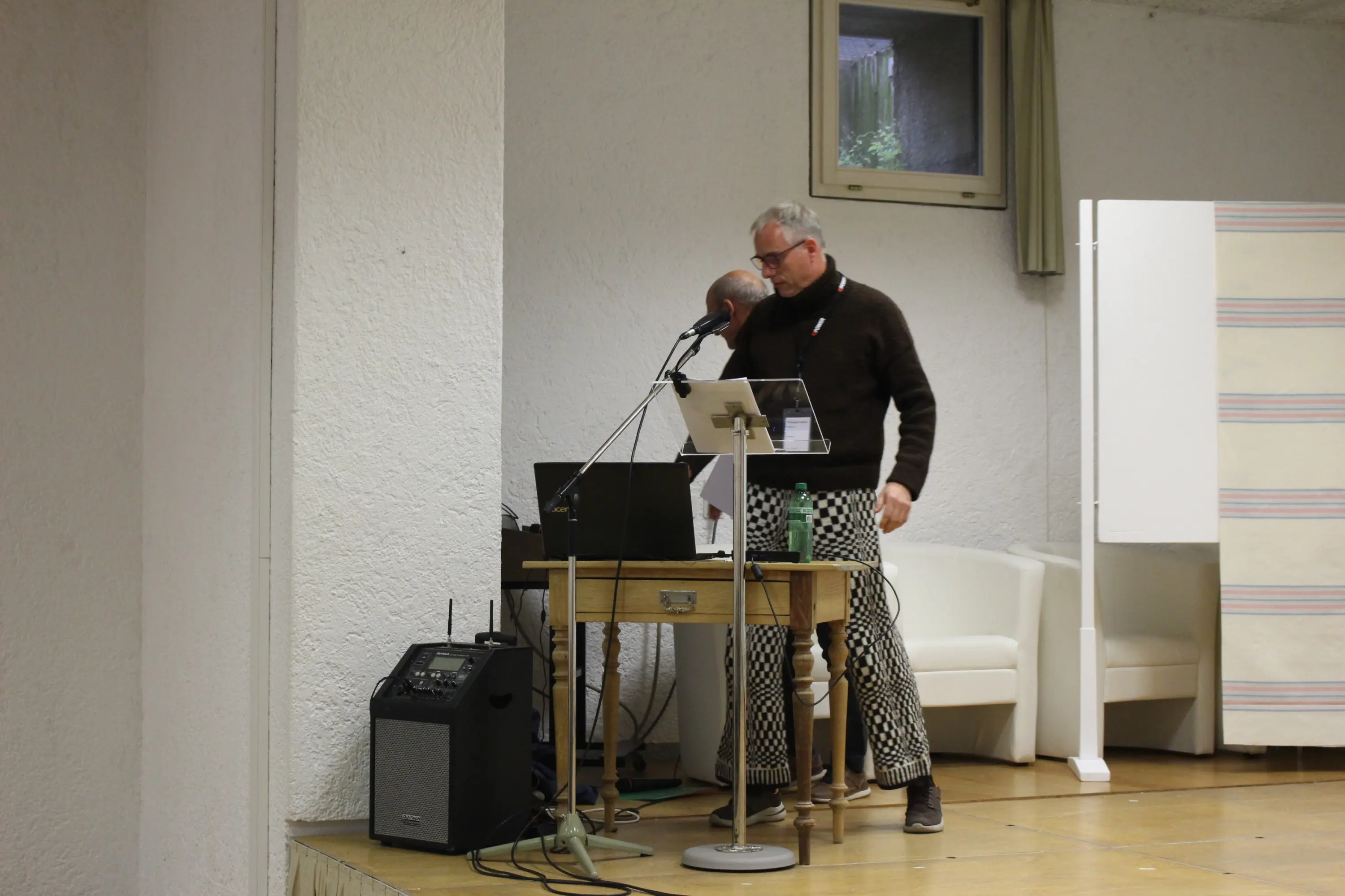 Un uomo con occhiali e maglione marrone si trova accanto a un tavolo con attrezzature audio. Sul tavolo ci sono un computer portatile e un microfono. Sullo sfondo, una parete bianca e una sedia bianca.