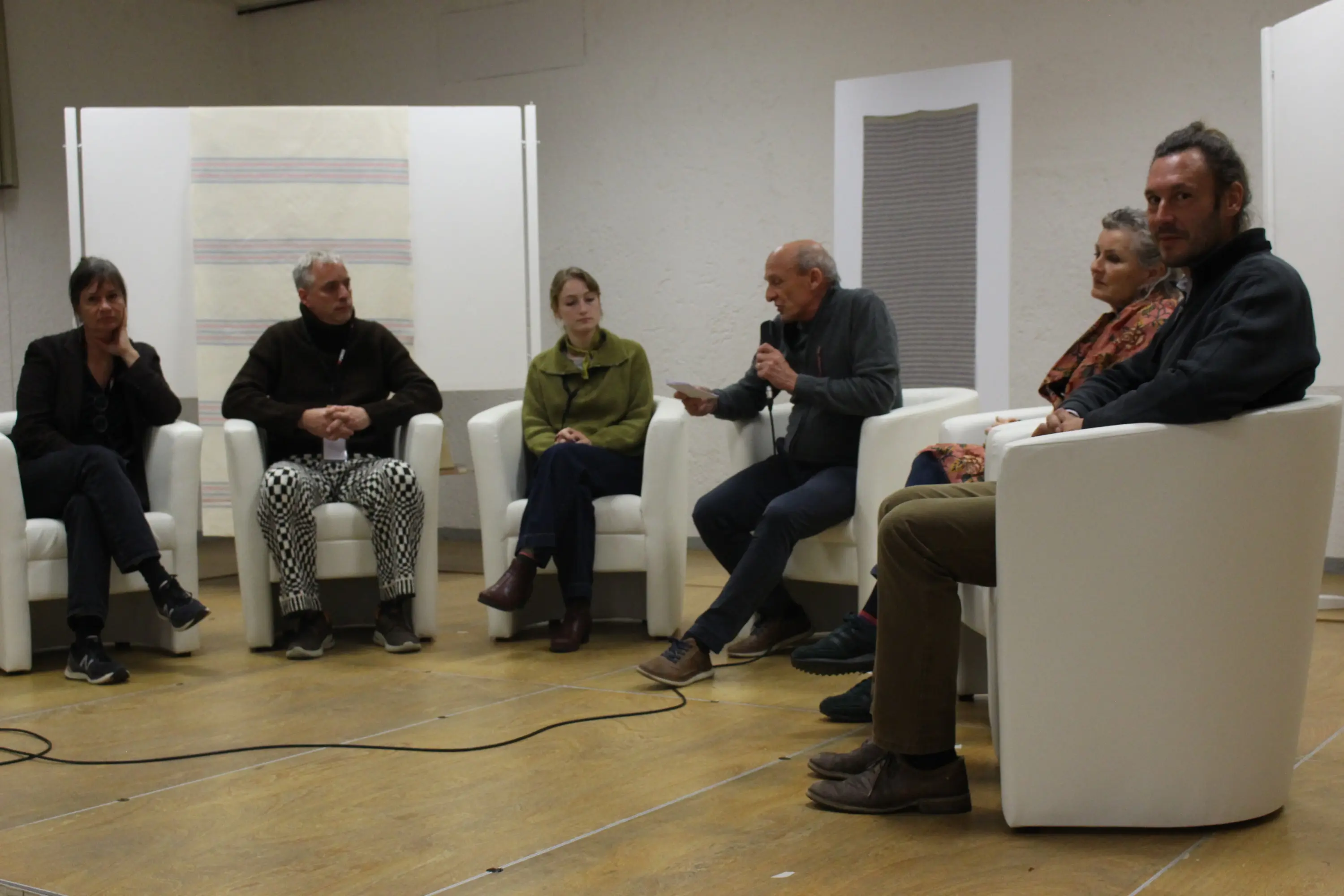 Cinque persone siedono su poltrone bianche in una sala. Un uomo, al centro, parla mentre gli altri ascoltano attentamente. L'ambiente è semplice e sobrio, con un parquet chiaro e pannelli dietro di loro.