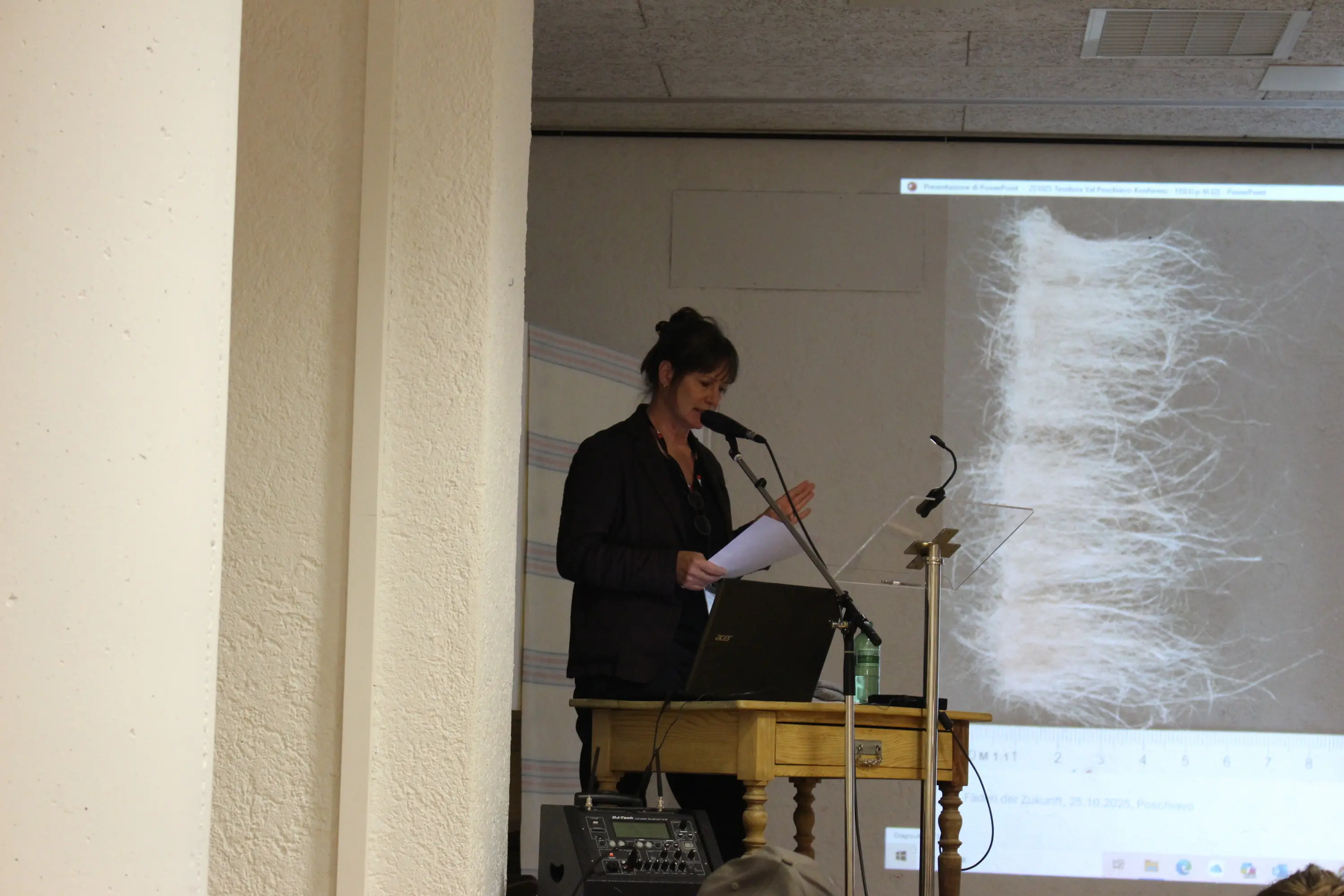 Una donna parla al microfono davanti a un tavolo con un computer portatile e una bottiglia d'acqua. Sullo sfondo, una proiezione mostra un'immagine di fibre.