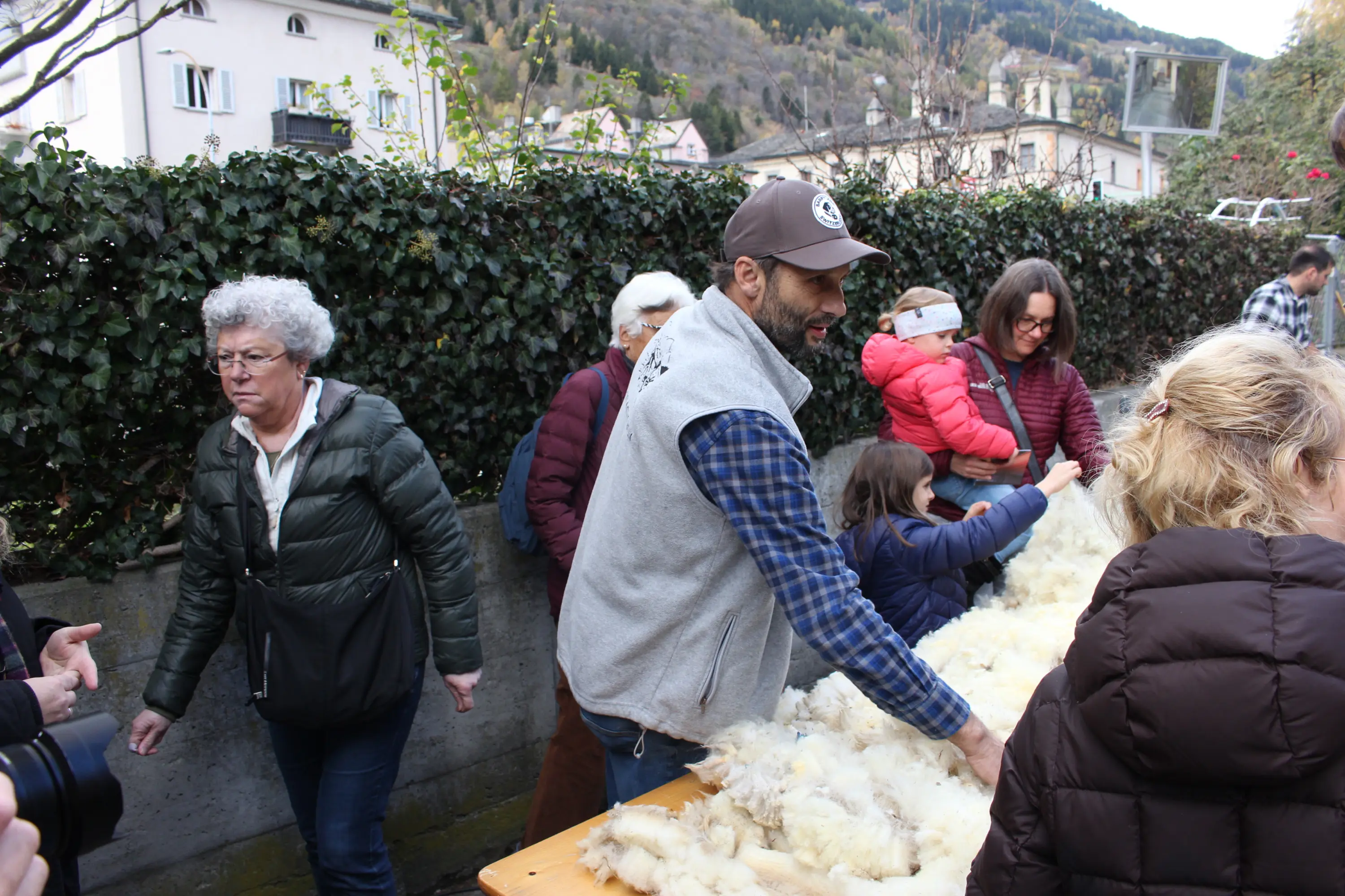 Persone di diverse età partecipano a un'attività all'aperto, lavorando insieme sulla lana. Un uomo in giacca grigia e cappellino aiuta una bambina, mentre altre persone osservano e interagiscono. Sullo sfondo, si intravedono case e