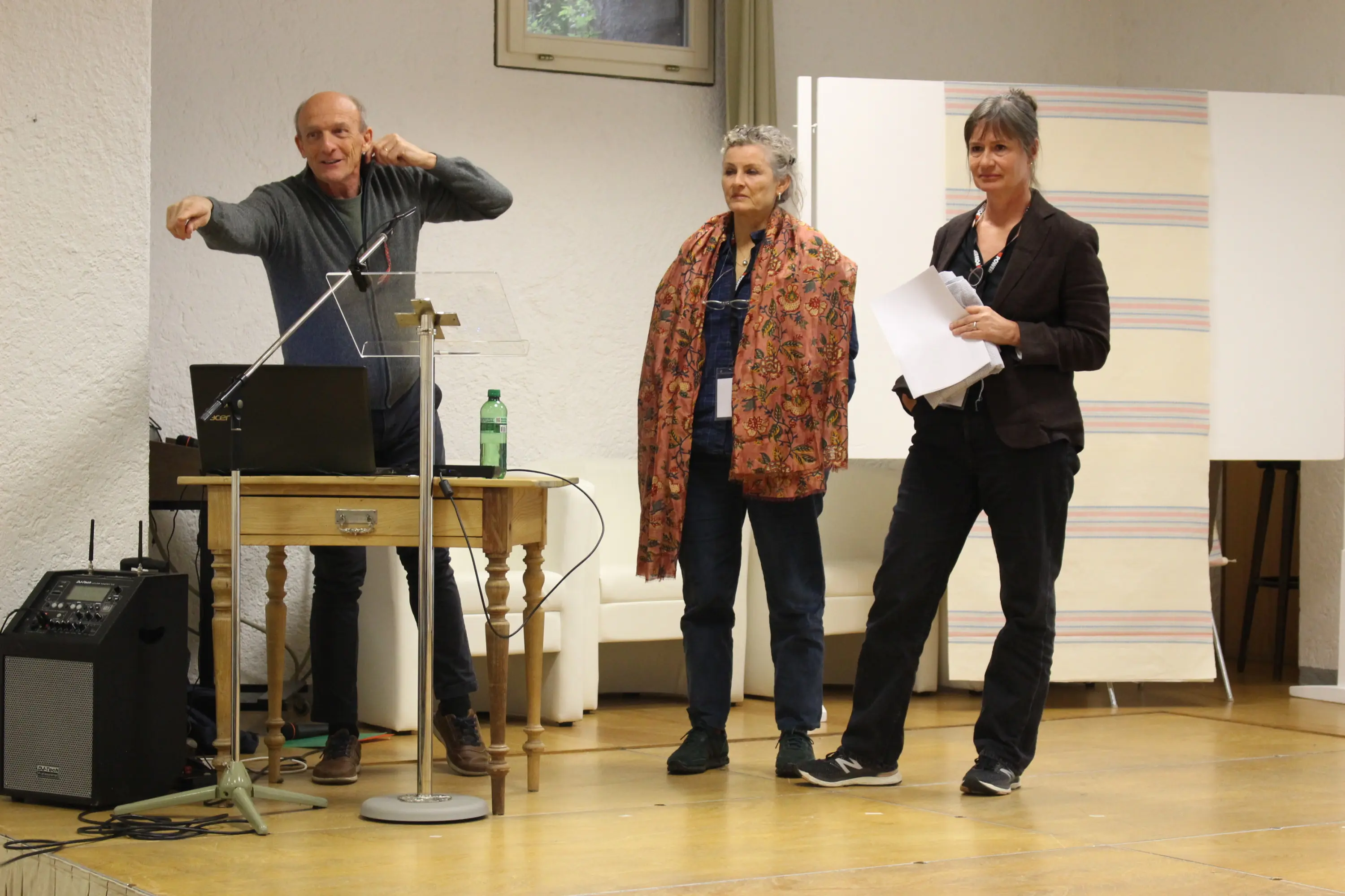 Tre persone su un palco durante una presentazione. Un uomo a sinistra, appassionato, gesticola mentre parla in un microfono. Due donne, una al centro con capelli ricci e una sciarpa colorata, e l'altra a destra con un documento in mano,