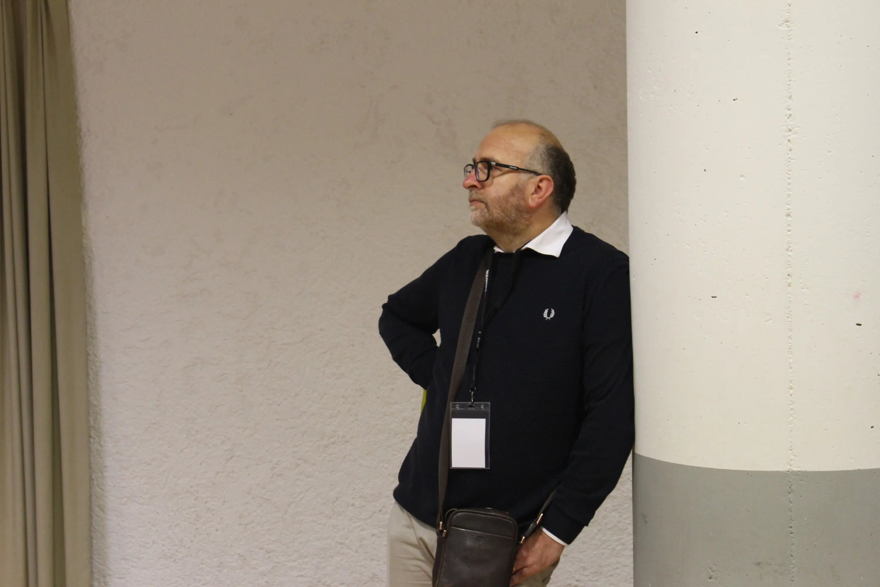 Un uomo in piedi accanto a un pilastro, con le braccia incrociate e un'espressione pensierosa. Indossa un maglione scuro e occhiali.