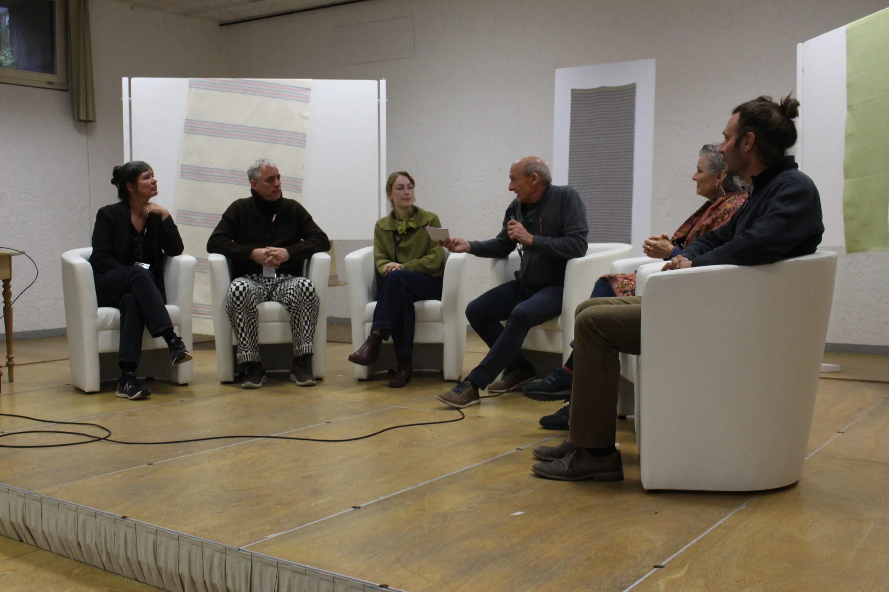 Un gruppo di sei persone si trova su un palco, sedute su poltrone bianche. Ognuno partecipa a una discussione, con un uomo anziano al centro che parla. Tutti indossano abiti casual e le pareti dietro di loro sono decorate