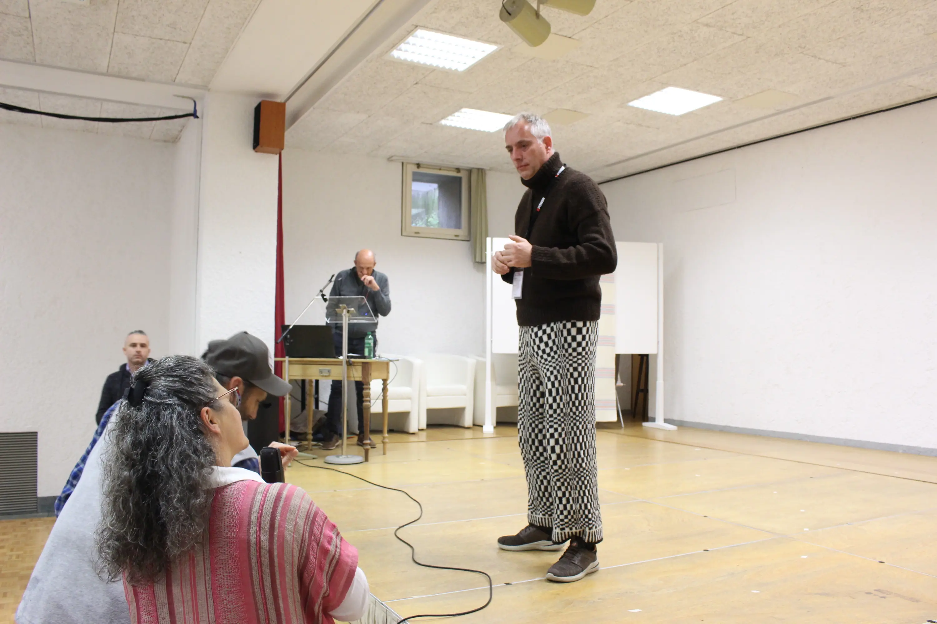 Un uomo in maglione scuro e pantaloni a scacchi parla a un pubblico seduto. Dietro di lui, c'è un uomo al computer e una sala con pareti chiare.