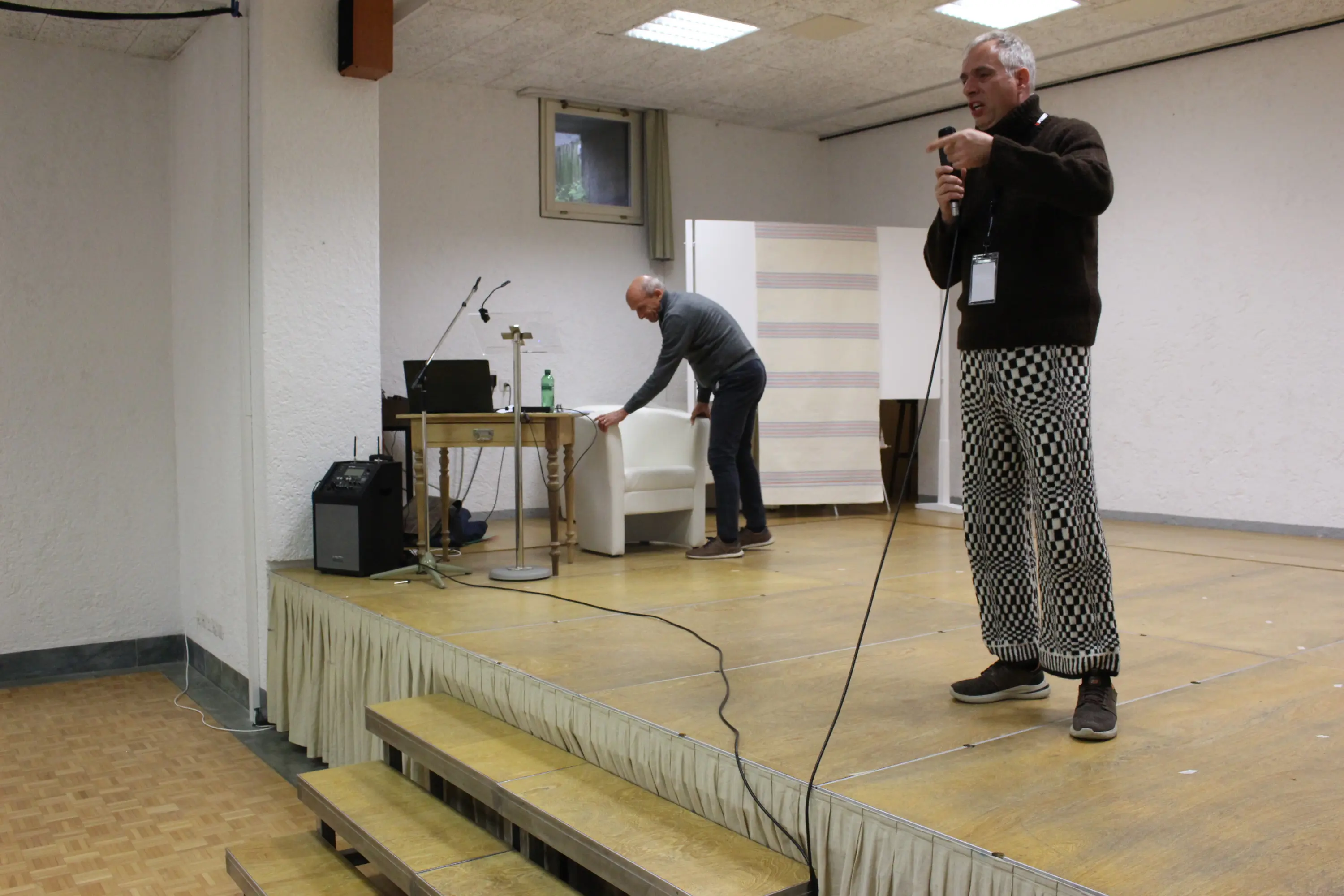 Una persona in piedi parla al microfono su un palco, mentre un'altra persona si sta preparando dietro di lui. Sul palco ci sono una sedia bianca e un tavolo con un computer portatile e una bottiglia d'acqua.