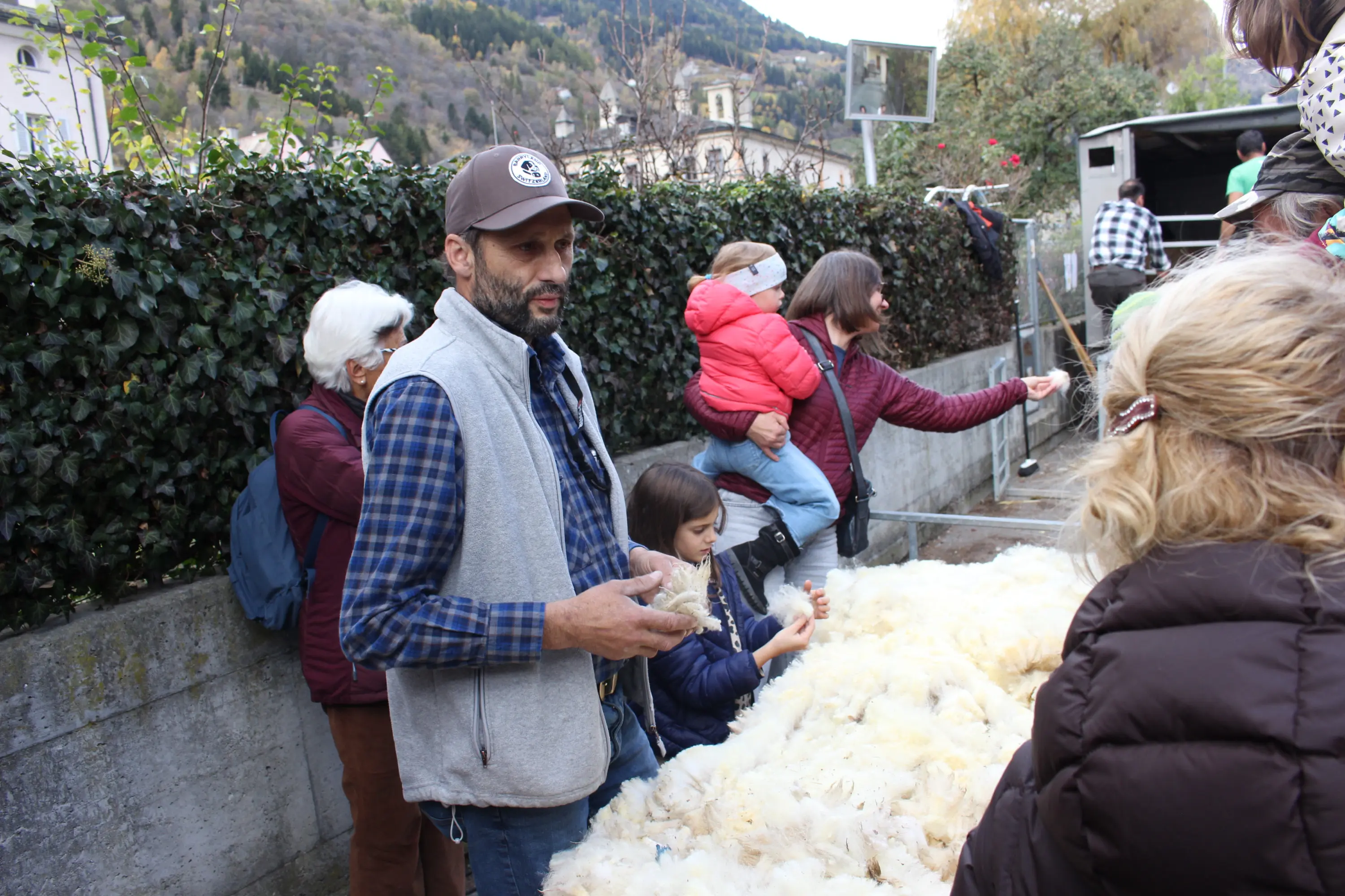 Un uomo con un cappellino e una giacca grigia parla mentre alcune persone, incluse due donne e bambini, si trovano intorno a un mucchio di ovatta. Sullo sfondo si vedono alberi e un paesaggio montano.