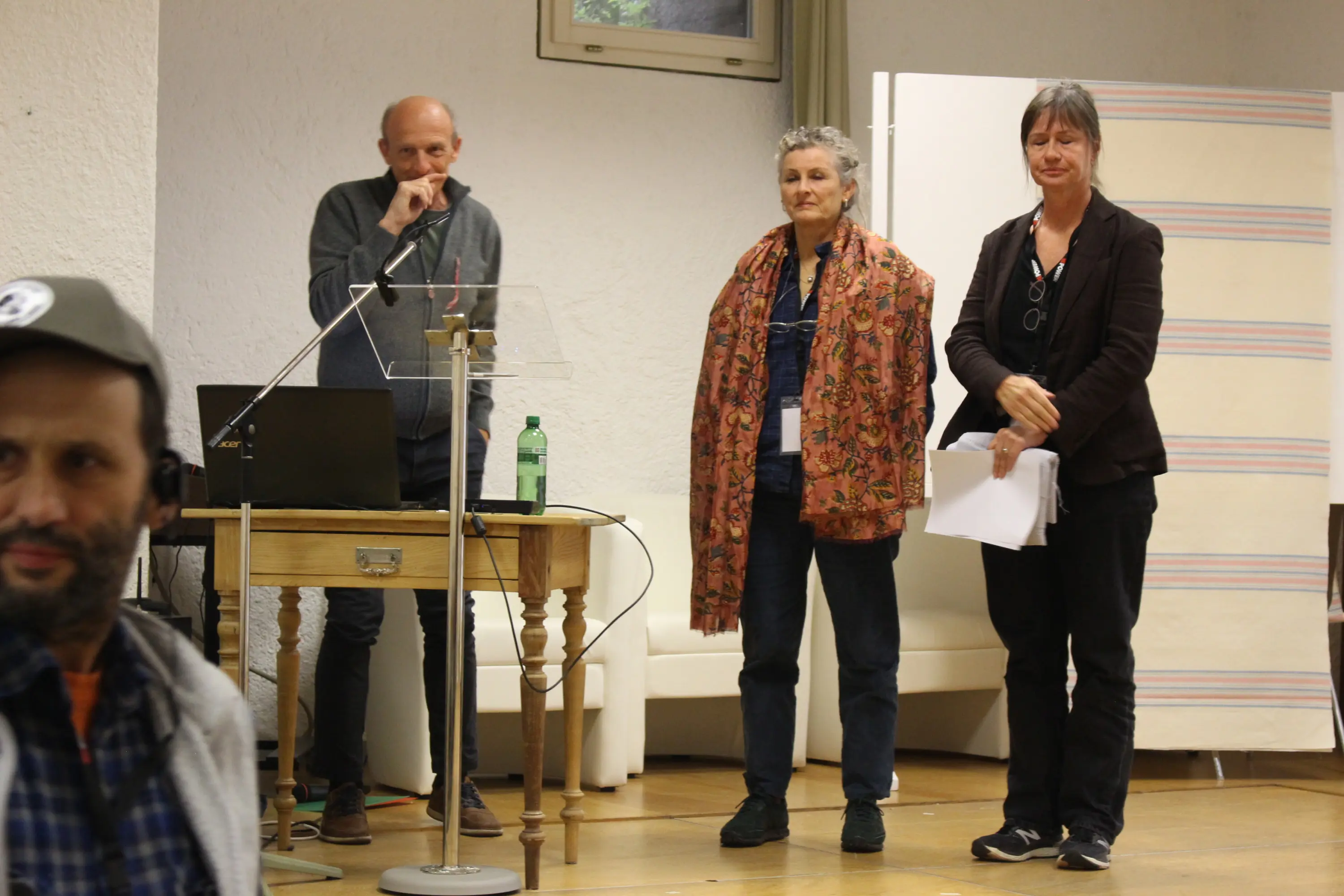 Tre persone stanno sul palco di un evento. A sinistra, un uomo con una maglietta grigia al microfono. Al centro, una donna con un grande scialle colorato. A destra, una donna con i capelli raccolti e una giacca scura. Un tav