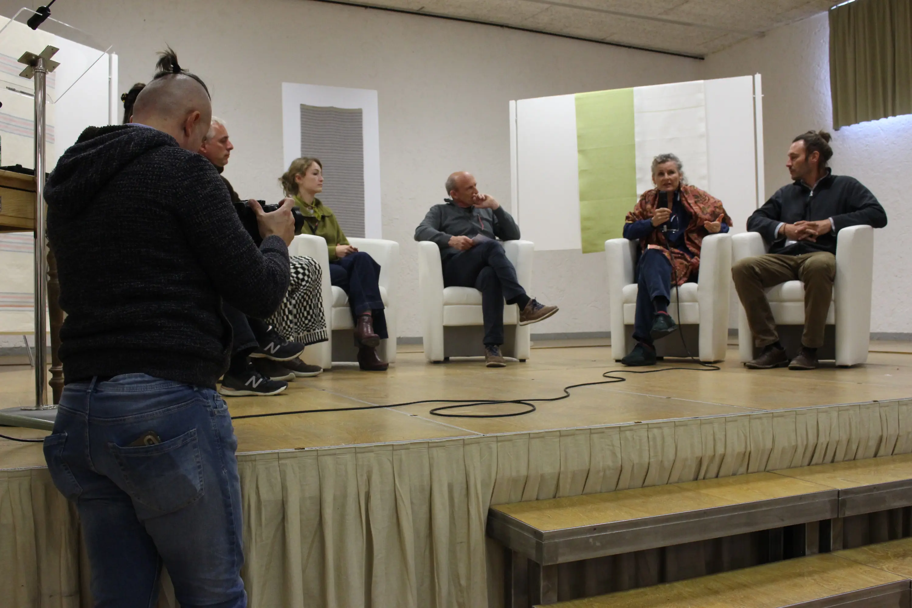 Un gruppo di cinque persone sedute su poltrone bianche durante un evento di discussione. Un fotografo è in primo piano, intento a scattare una foto. Sullo sfondo si vede un palco semplice con decorazioni minimaliste.
