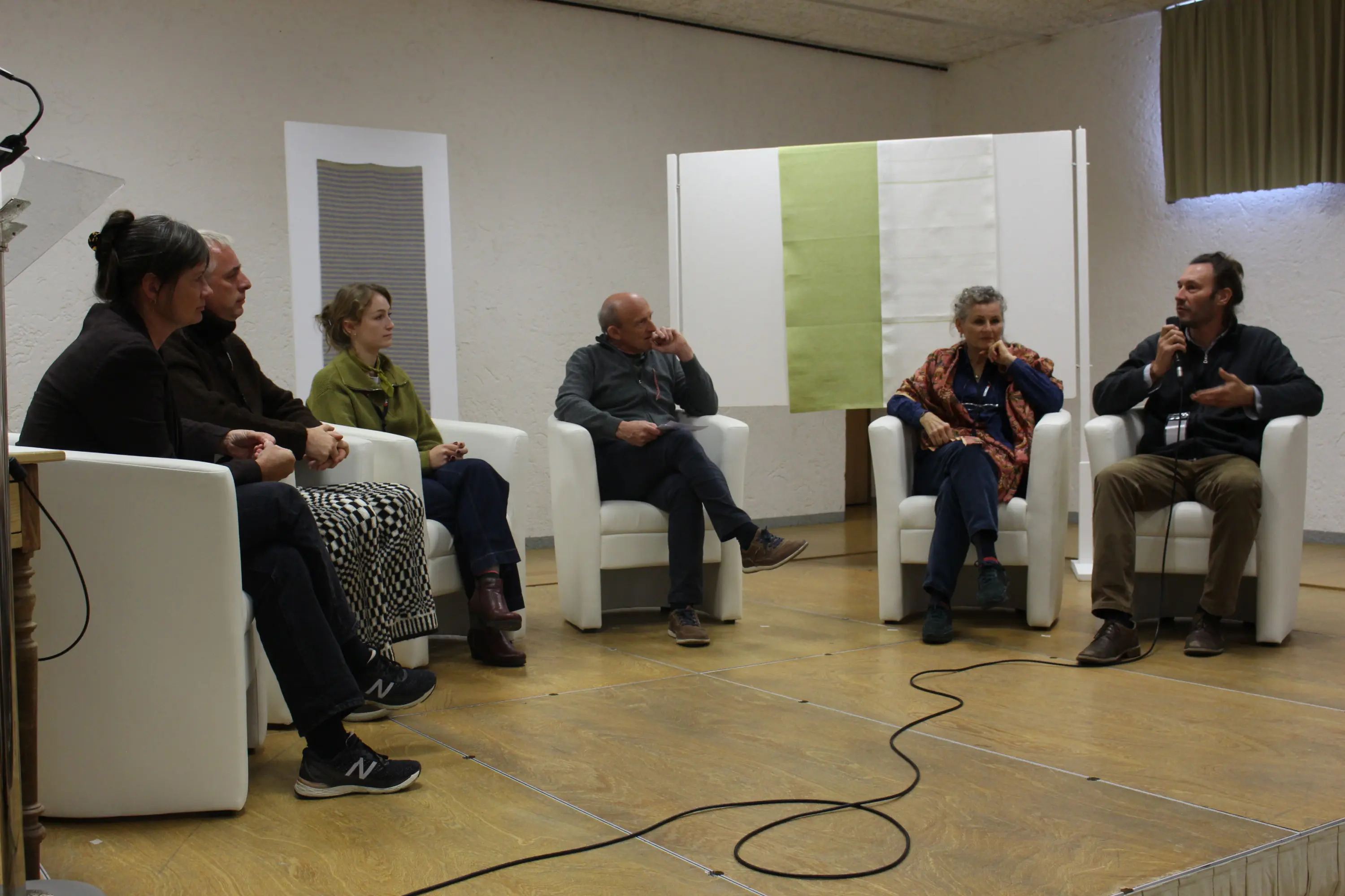 Un gruppo di sei persone sedute su poltrone bianche in un ambiente interno. Sono coinvolti in una discussione, con uno dei partecipanti che parla mentre gli altri ascoltano attentamente. Sullo sfondo, un pannello bianco e verde illumina la scena.