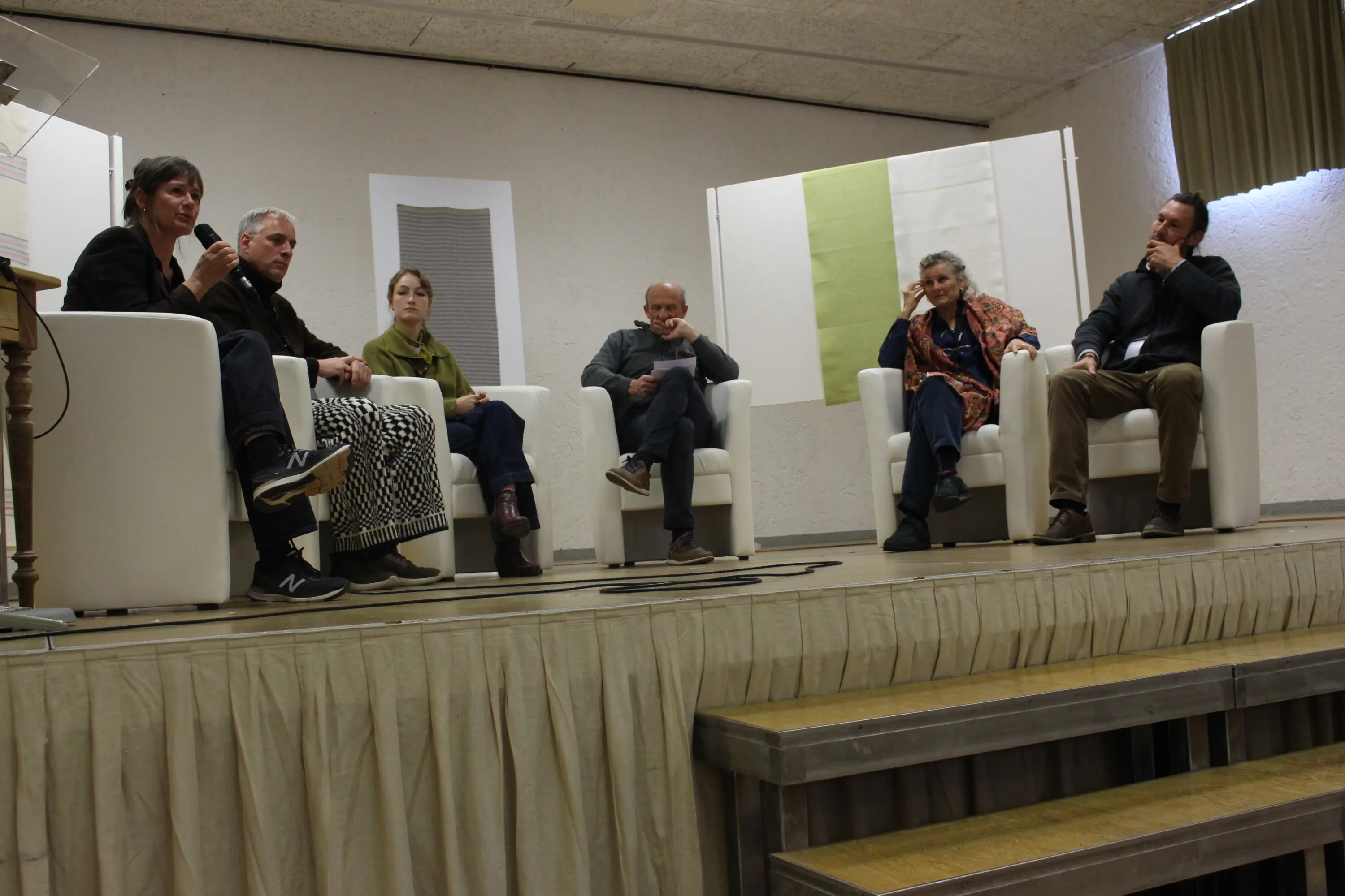 Un gruppo di sei persone sedute su poltrone bianche durante una discussione su un palco. Un relatrice parla mentre gli altri ascoltano con interesse. Sono presenti diverse espressioni e abbigliamenti. Sullo sfondo, ci sono pannelli con texture o colori