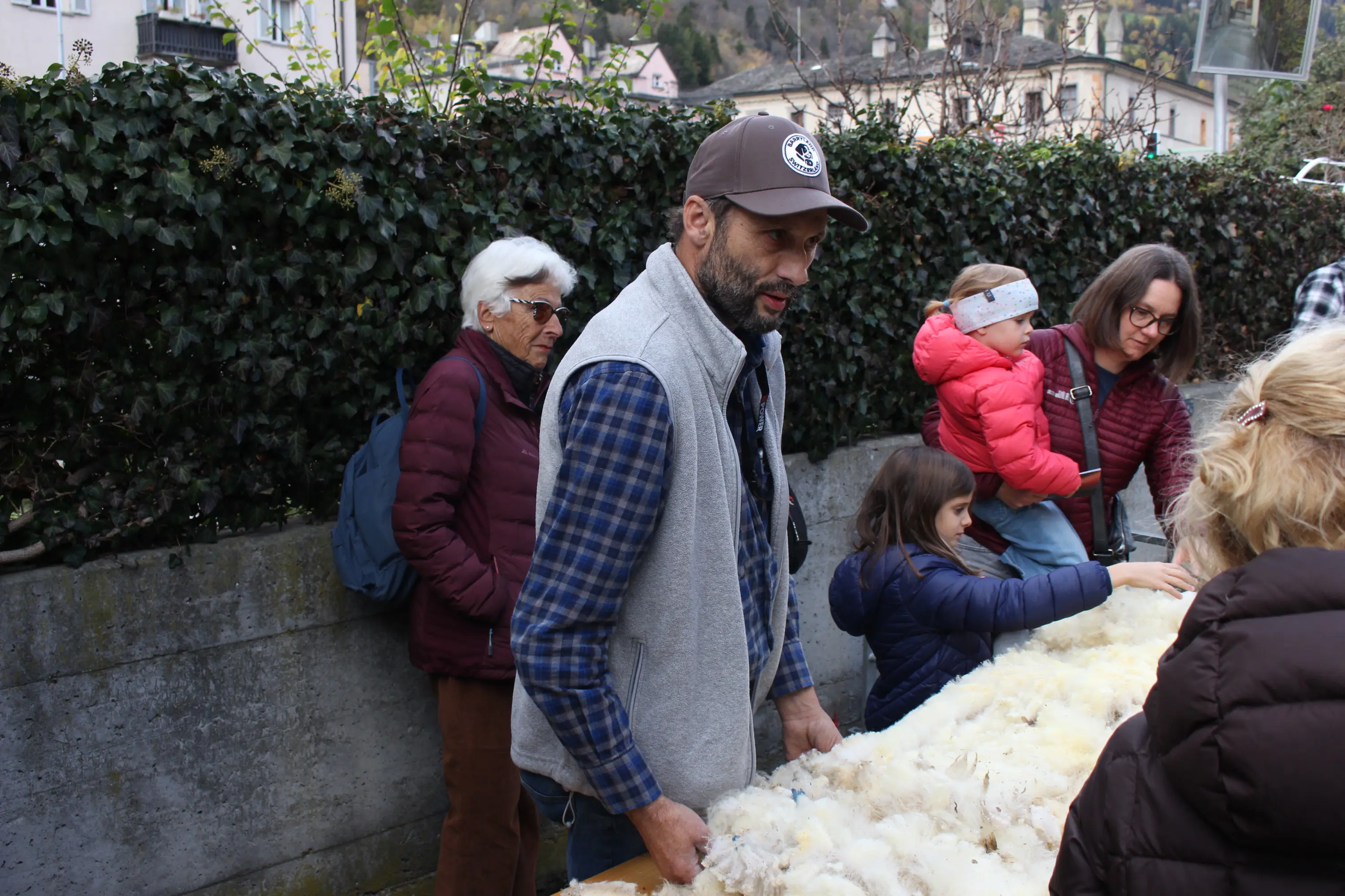 Un gruppo di persone in una fiera all'aperto, con un uomo che manipola un tappeto di piume. Alcuni partecipanti osservano, inclusa una bambina con una giacca blu e una donna che tiene un bambino. Sullo sfondo si intravedono edifici