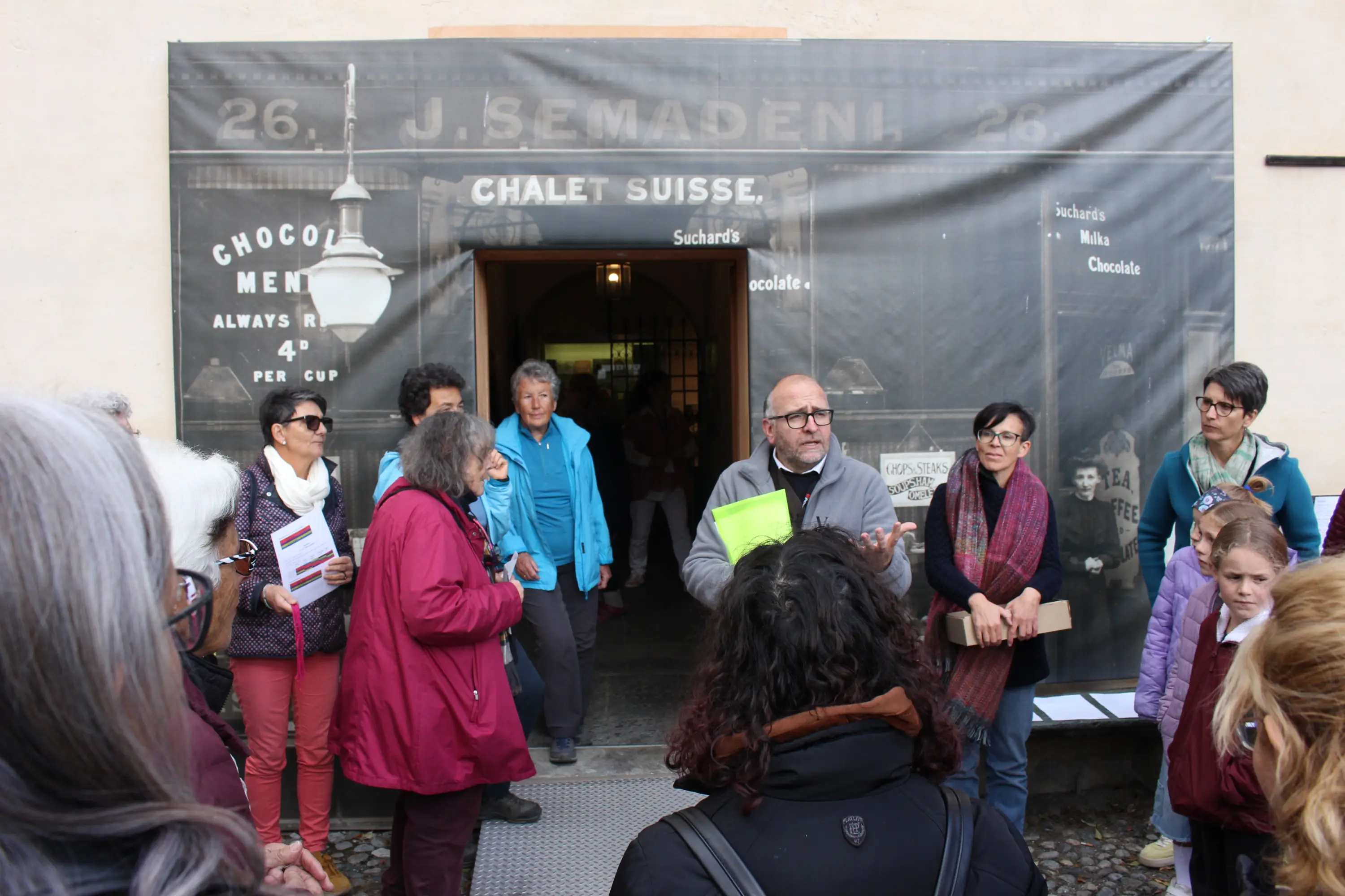 Un gruppo di persone di diverse età si riunisce davanti a una porta d’ingresso. Un uomo, con occhiali e una cartella, parla al gruppo che ascolta attentamente. Sullo sfondo, un grande striscione pubblicitario di un vecchio negozio di