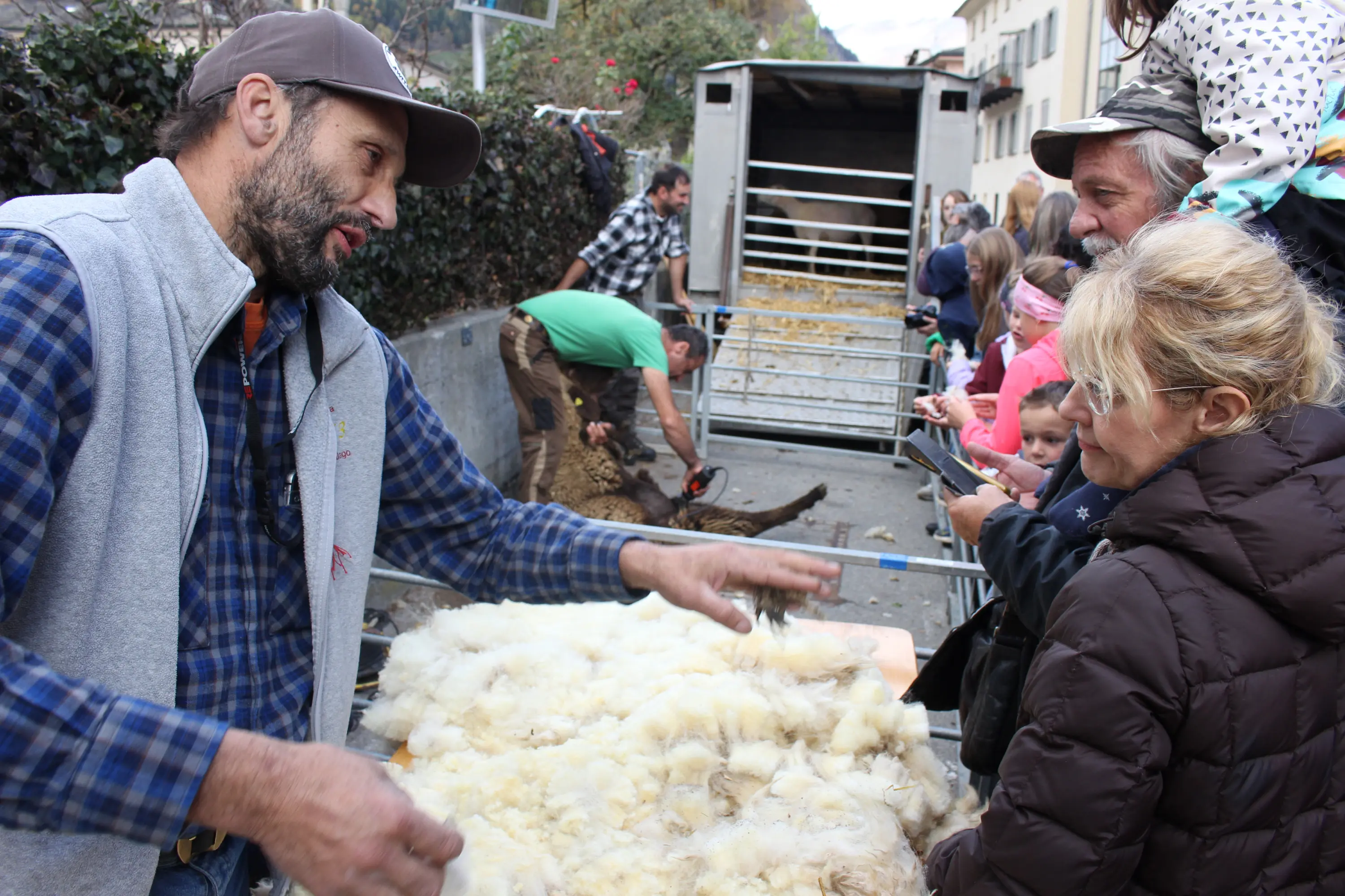 Un allevatore mostra della lana a un gruppo di persone durante un evento all'aperto. Sullo sfondo, altri partecipanti sono impegnati in attività legate all'allevamento.