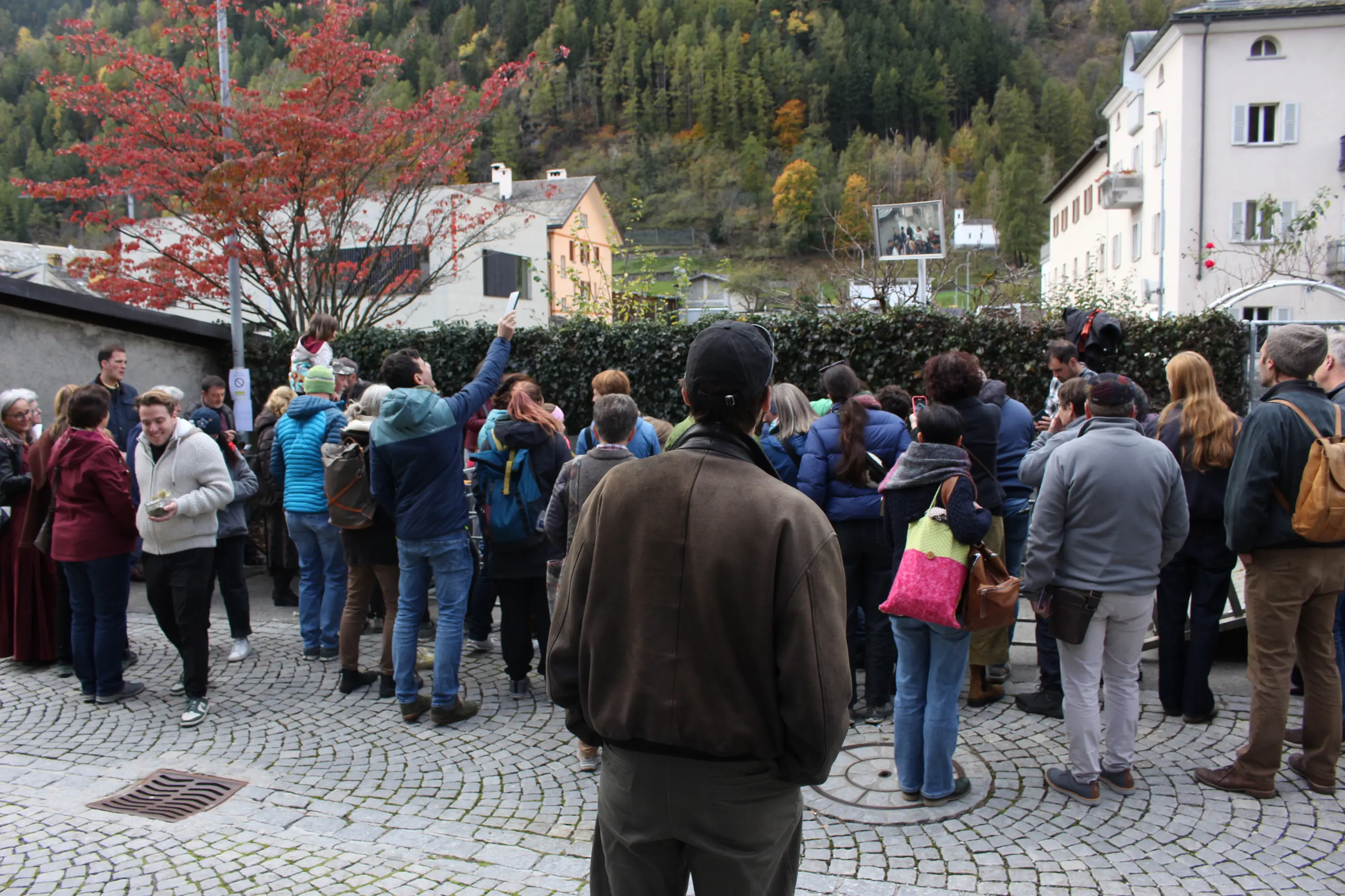 Un gruppo di persone si raduna in una piazza, alcune sollevano le mani mentre altre osservano. Dietro di loro c'è un uomo in primo piano che guarda la scena. Sullo sfondo, edifici e alberi autunnali.