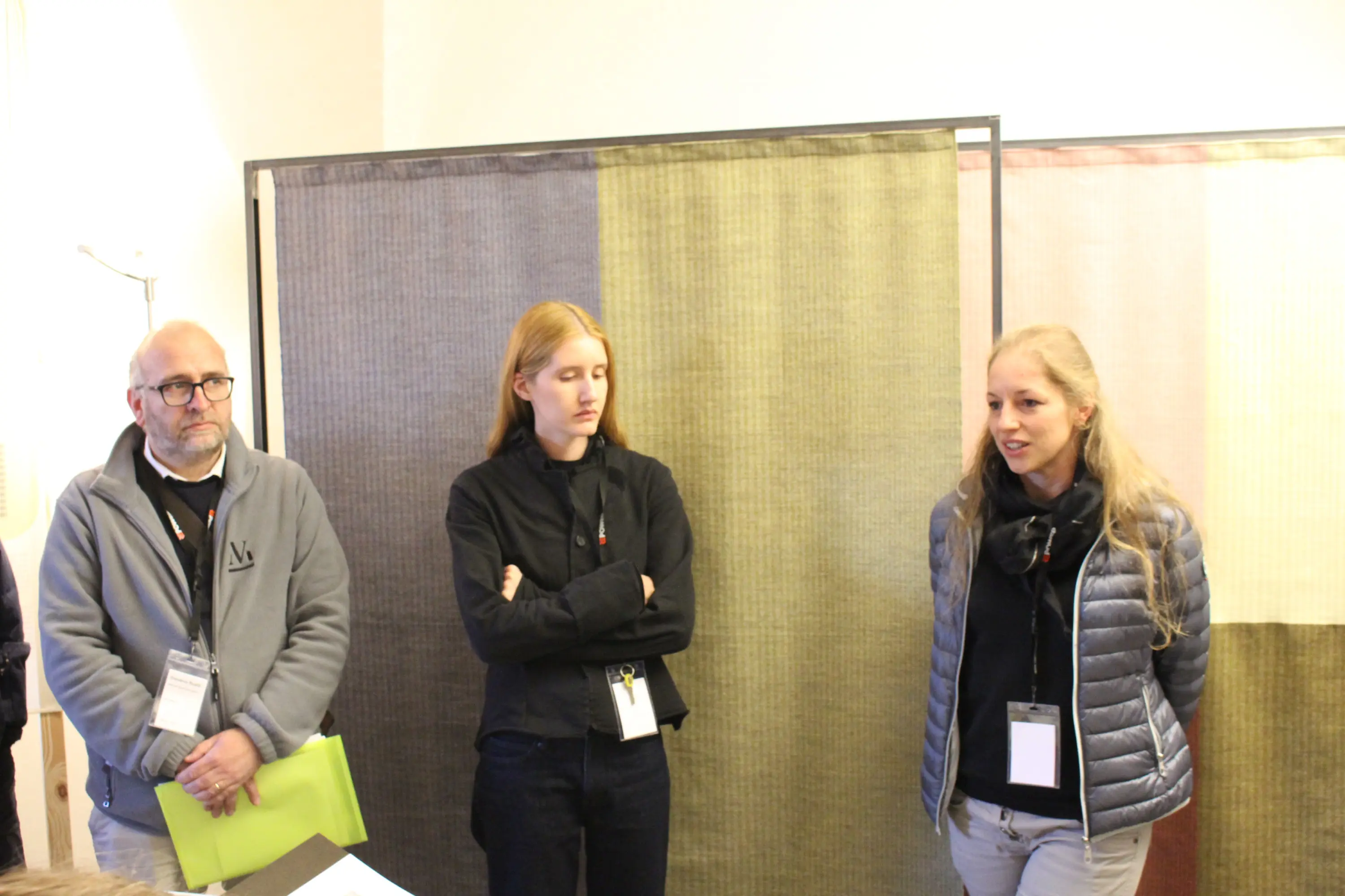 Tre persone stanno partecipando a una discussione. Due di loro, un uomo con occhiali e una donna con capelli lunghi e biondi, ascoltano attentamente mentre una donna con capelli lunghi e chiari sta parlando. Sullo sfondo si vedono pannelli