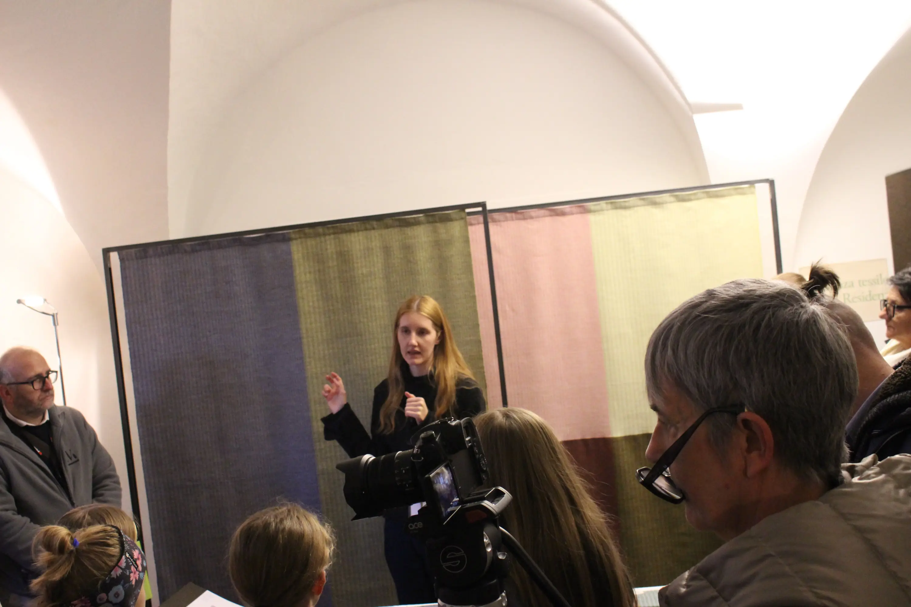 Una relatrice parla a un pubblico, con uno sfondo di tessuti colorati e una fotocamera in primo piano.