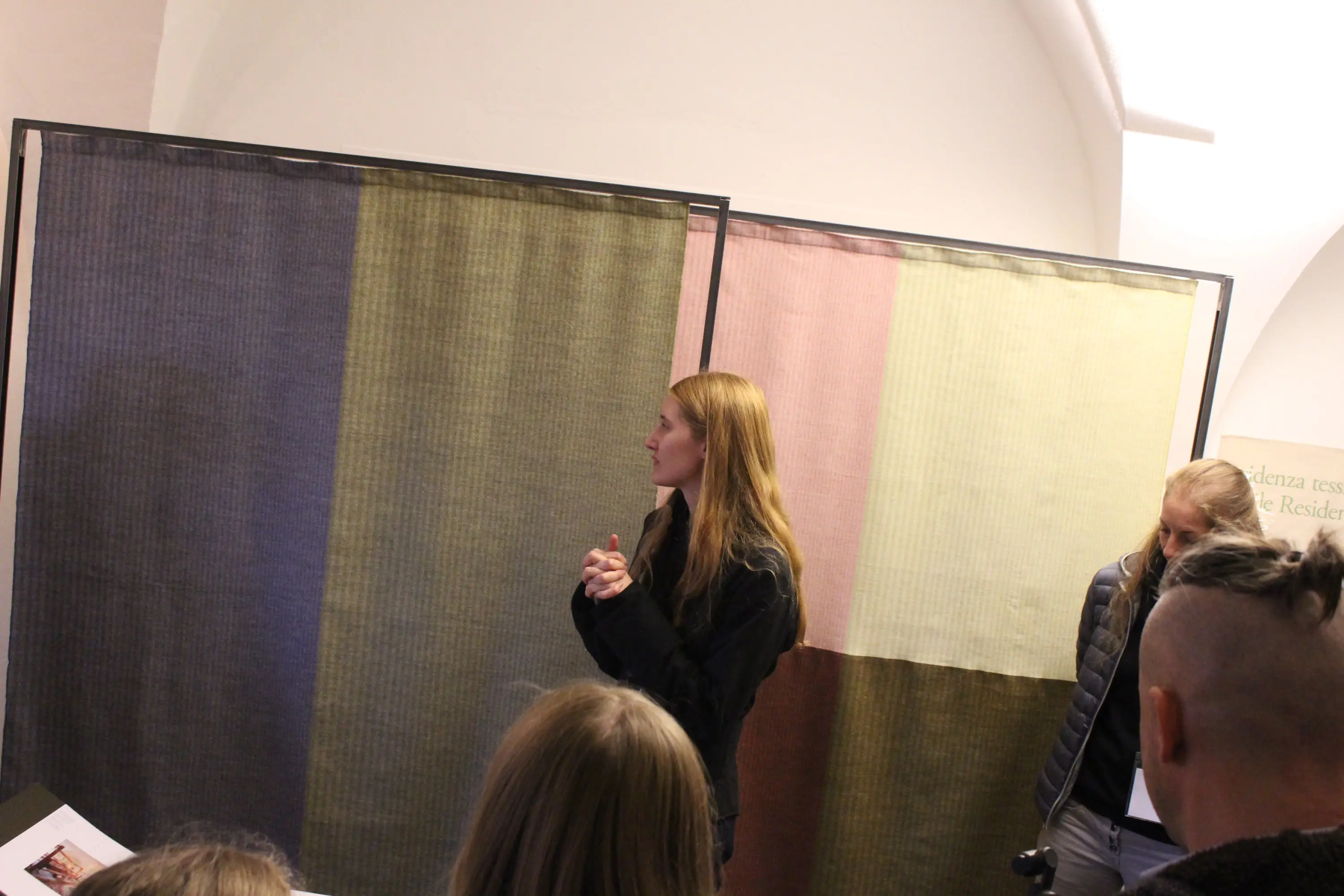 Una giovane donna con i capelli lunghi e biondi parla davanti a due grandi pannelli tessili di colori diversi, mentre un gruppo di persone ascolta attentamente.
