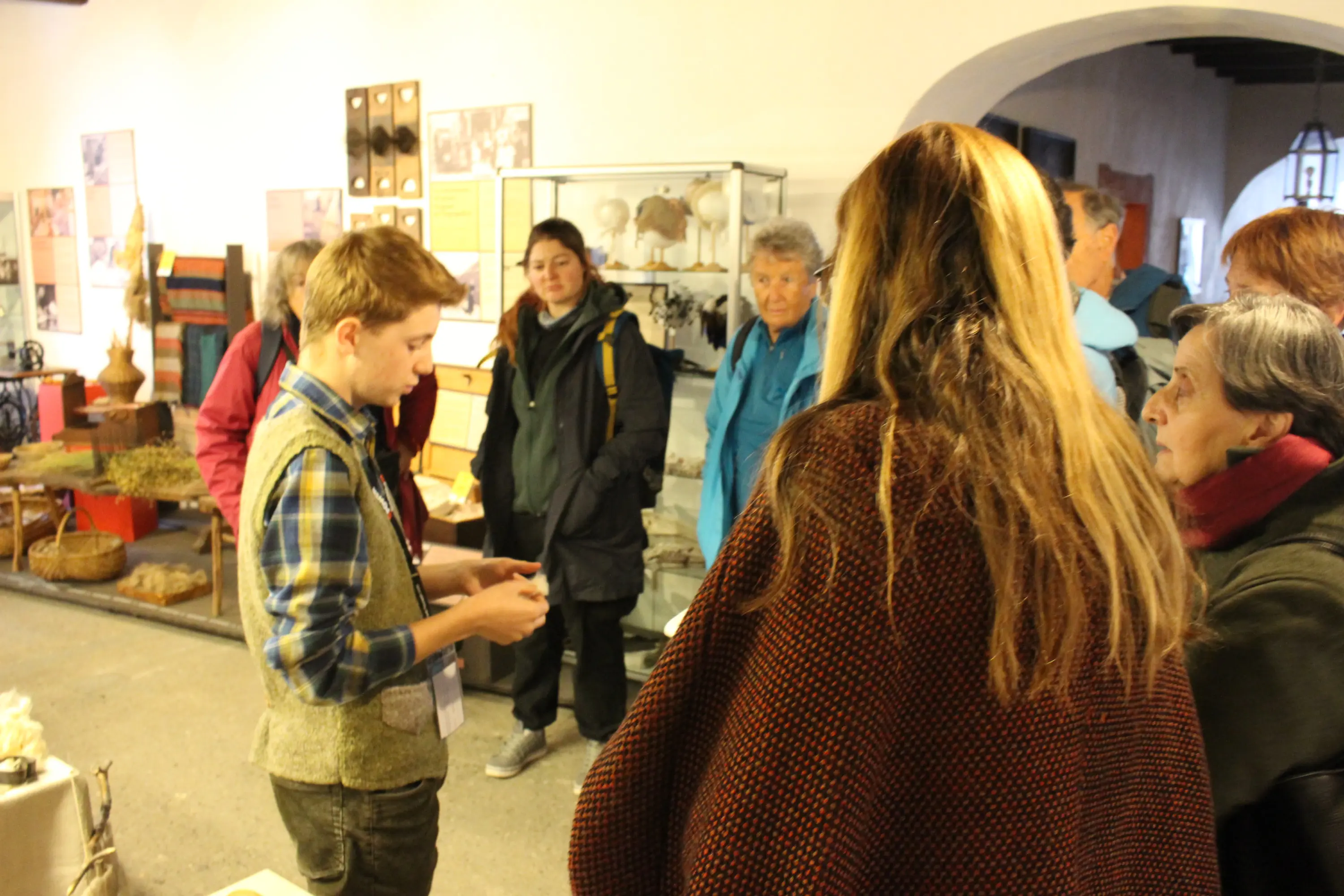 Un giovane guida mostra un oggetto a un gruppo di visitatori in un museo. Alcuni partecipanti osservano attentamente, mentre altri sono interessati alle esposizioni sullo sfondo.