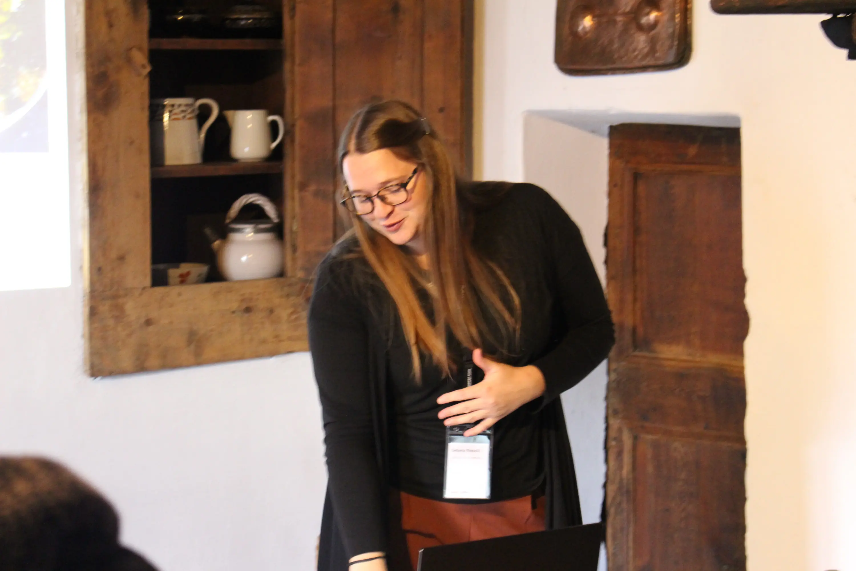 Una donna con lunghi capelli castani e occhiali sta presentando a un pubblico in un ambiente conviviale, con pareti di legno e arredi rustici. Sembra coinvolta e spiega qualcosa di interessante.