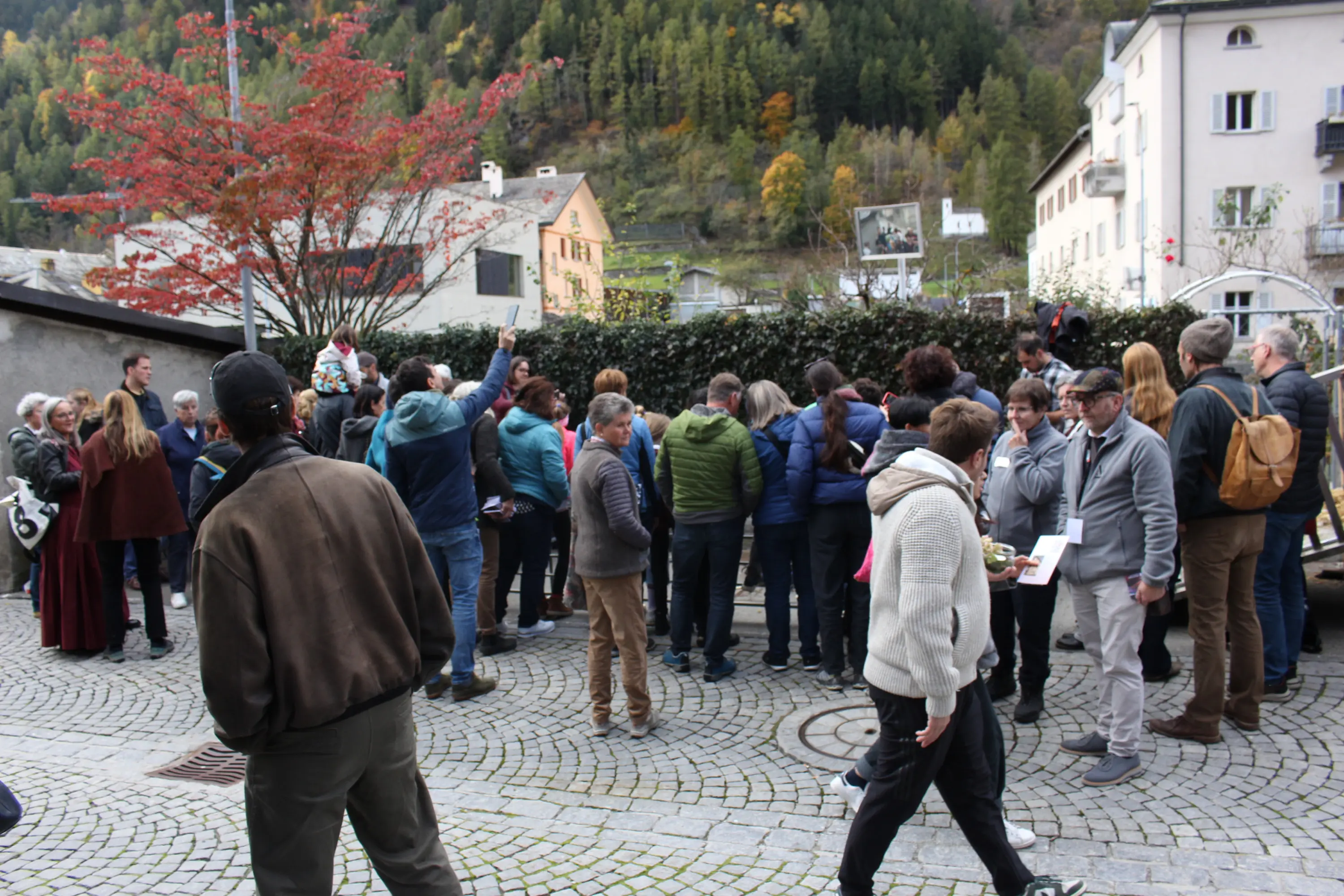 Un gruppo di persone si riunisce in una piazza circondata da alberi e case, con montagne sullo sfondo. Alcuni partecipanti indossano giacche colorate e stanno interagendo tra di loro.