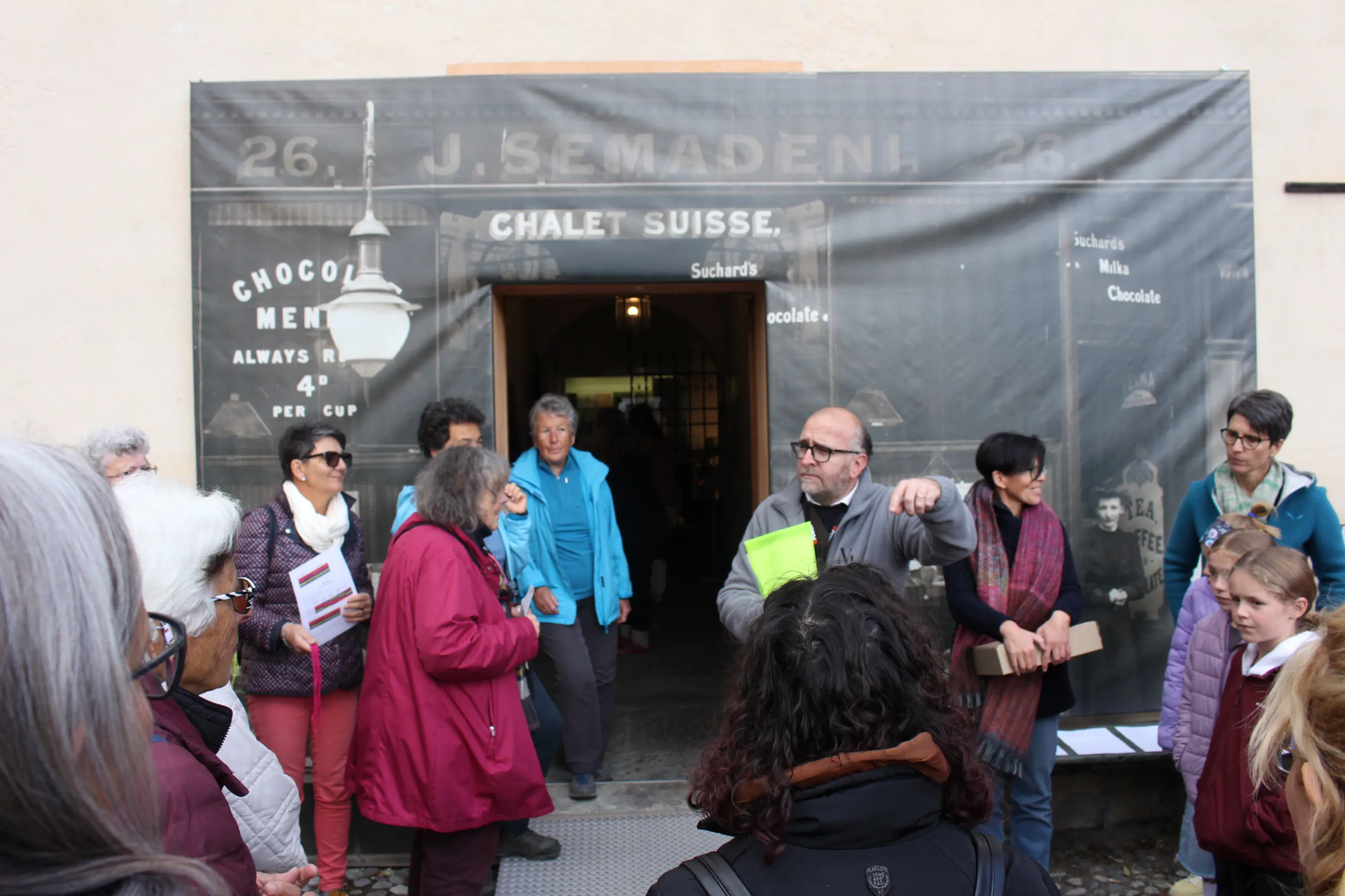 Gruppo di persone all'ingresso di un edificio con una facciata decorata da un striscione che riporta il nome "J. Semadeni" e riferimenti al cioccolato. Un uomo, probabilmente una guida, sta spiegando qualcosa al pubblico.
