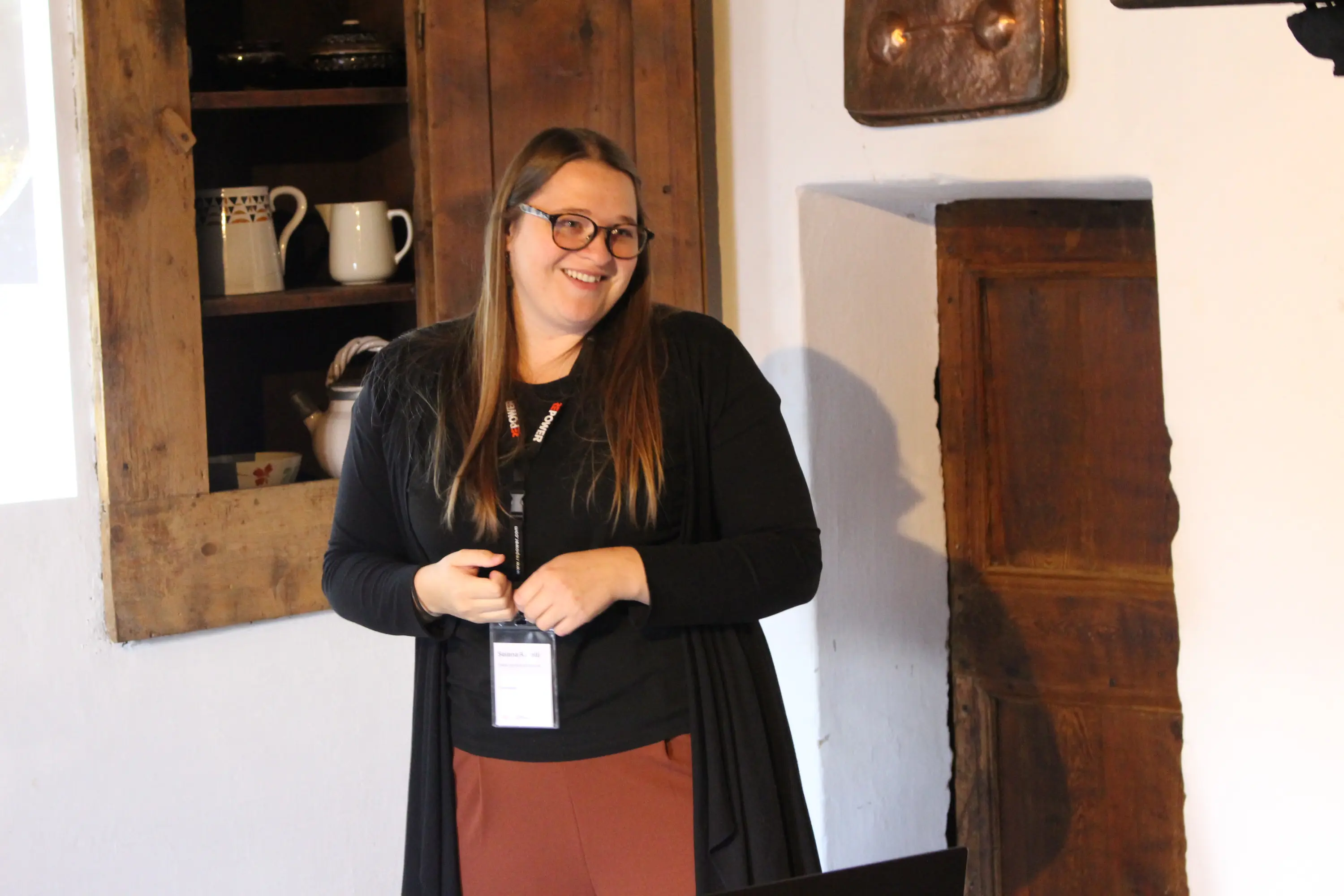 Una donna sorridente presenta in una stanza con pareti bianche e mobili in legno. Indossa una maglietta nera e un cardigan lungo, mentre tiene un microfono in mano.