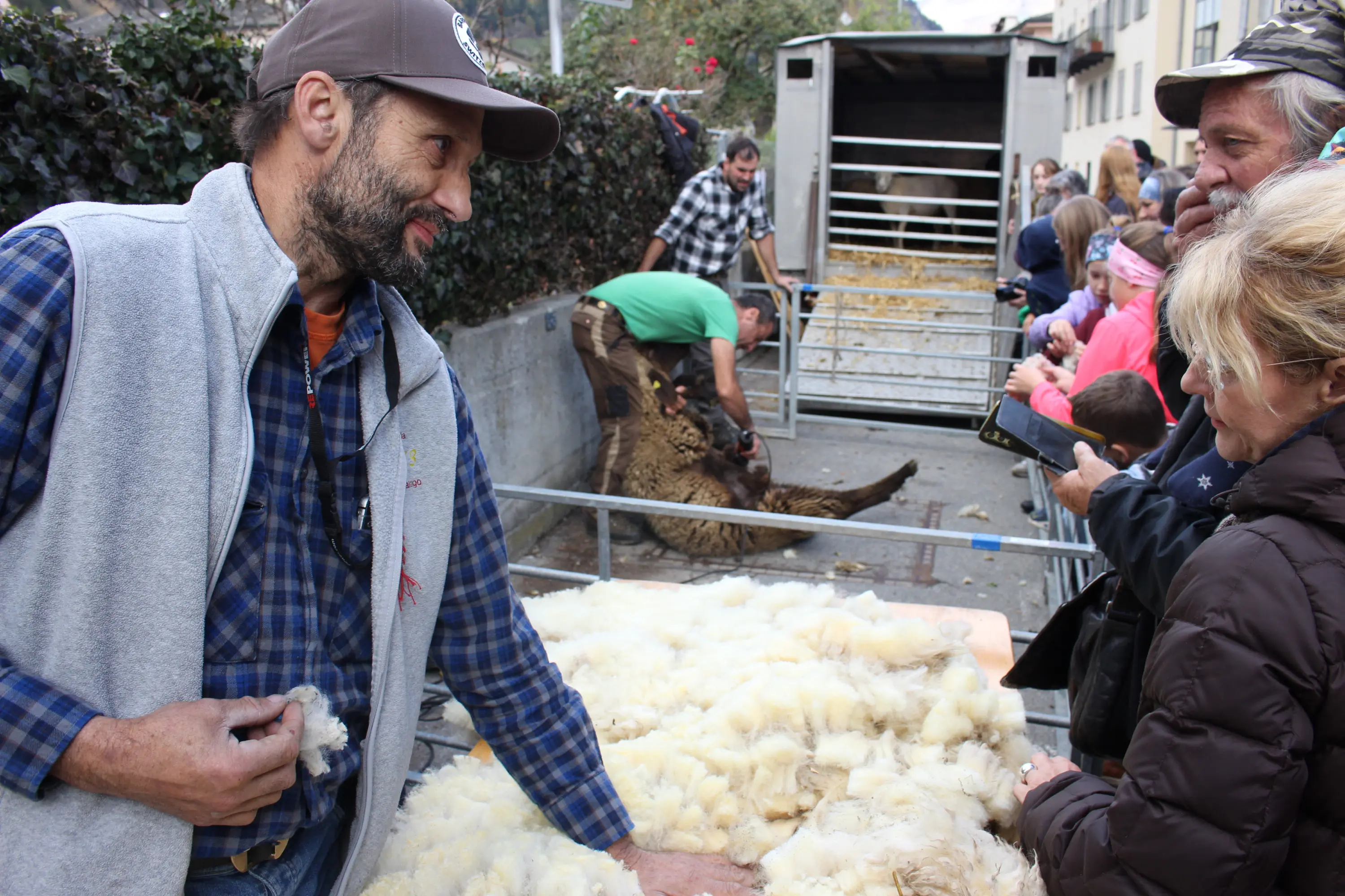 Un uomo sorride mentre mostra della lana a un gruppo di persone interessate. Sullo sfondo, un altro uomo si prende cura di una pecora, mentre alcuni bambini osservano con curiosità.