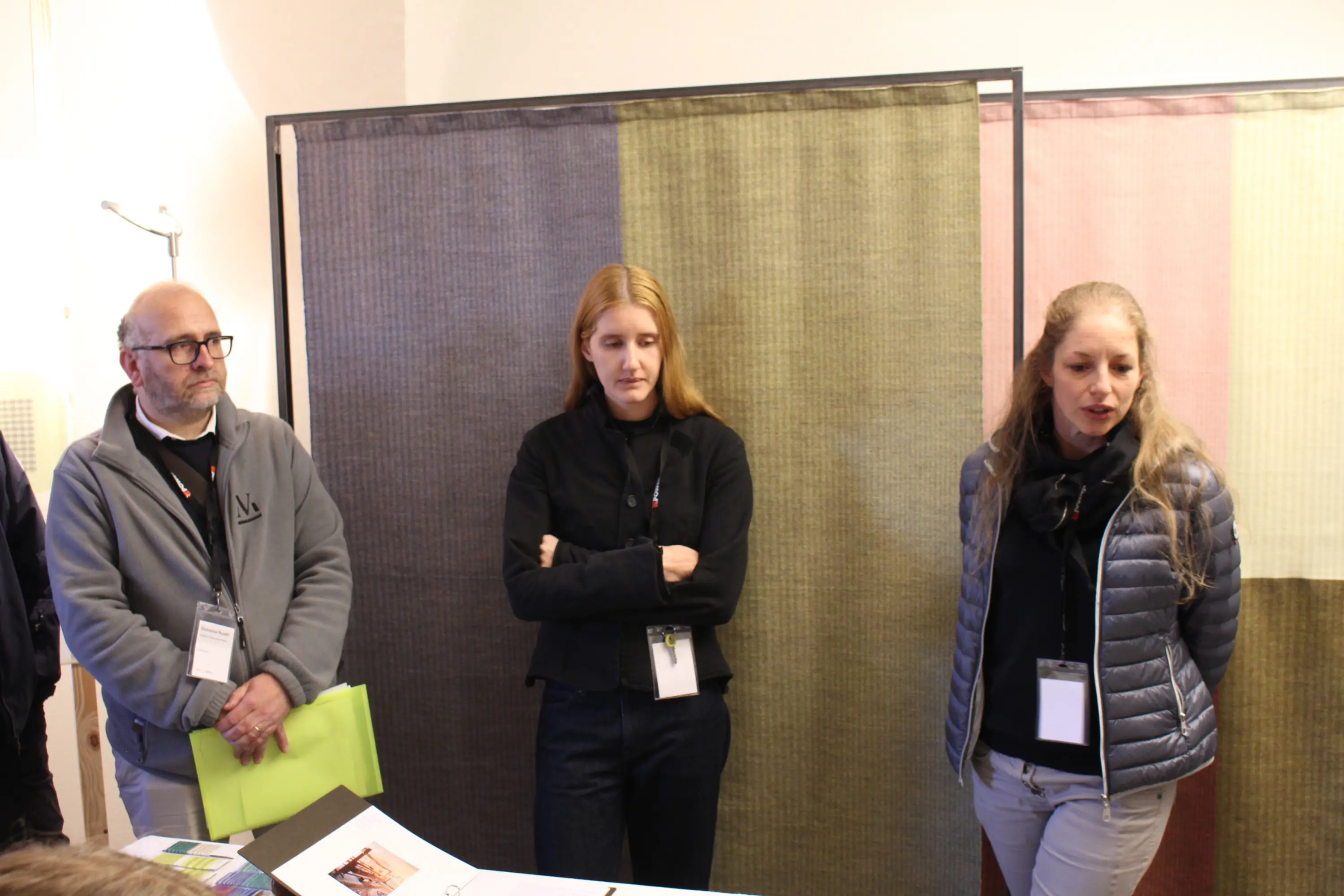 Tre persone in un ambiente interno, due donne e un uomo. La donna sulla destra parla, mentre gli altri due ascoltano. Sullo sfondo ci sono pannelli di tessuto colorato.