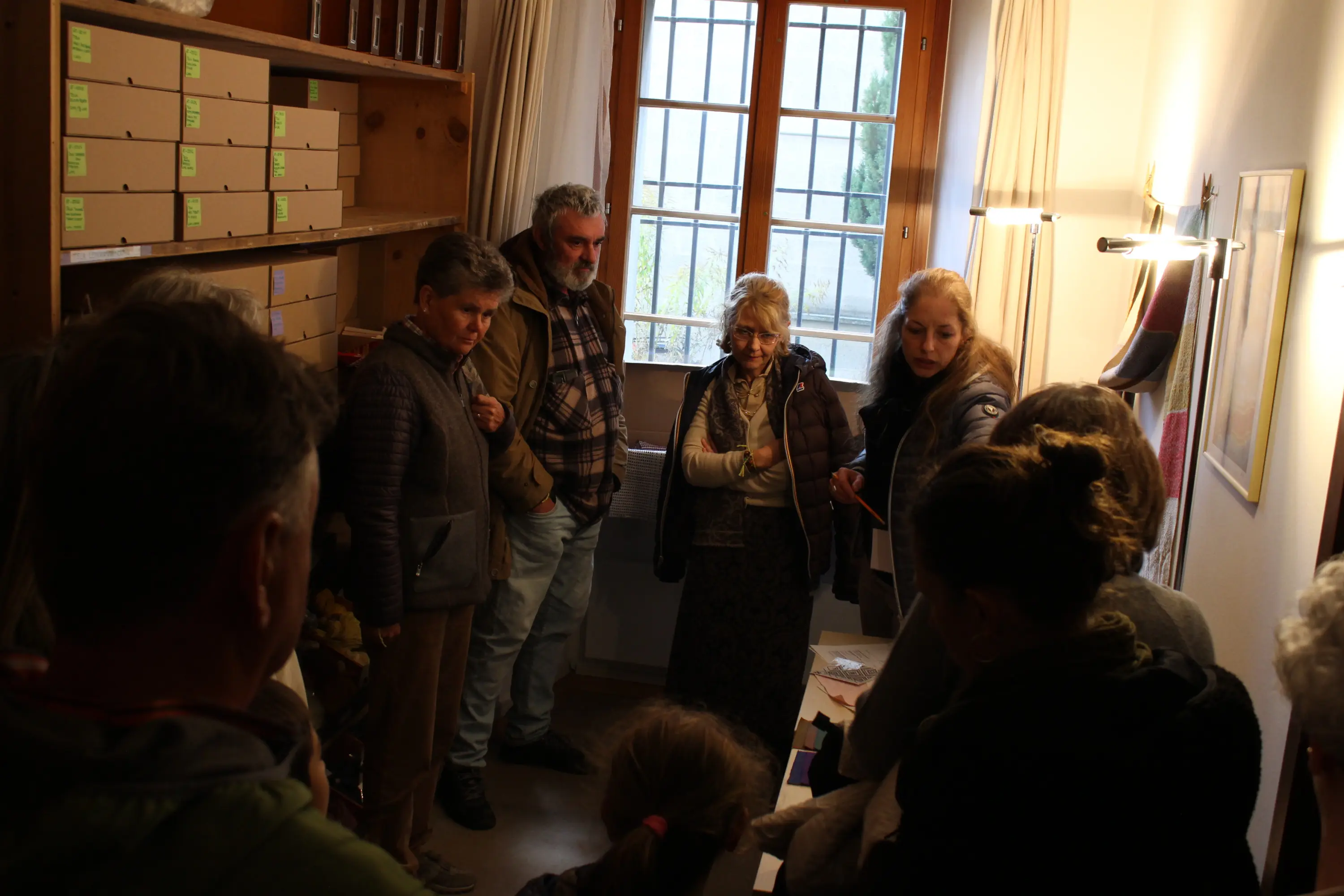 Un gruppo di persone si riunisce in una stanza con finestre, mentre ascoltano e discutono. Alcuni membri sembrano interessati e concentrati sulla conversazione. Le pareti sono decorate con articoli e ci sono scatole di cartone su uno scaffale.