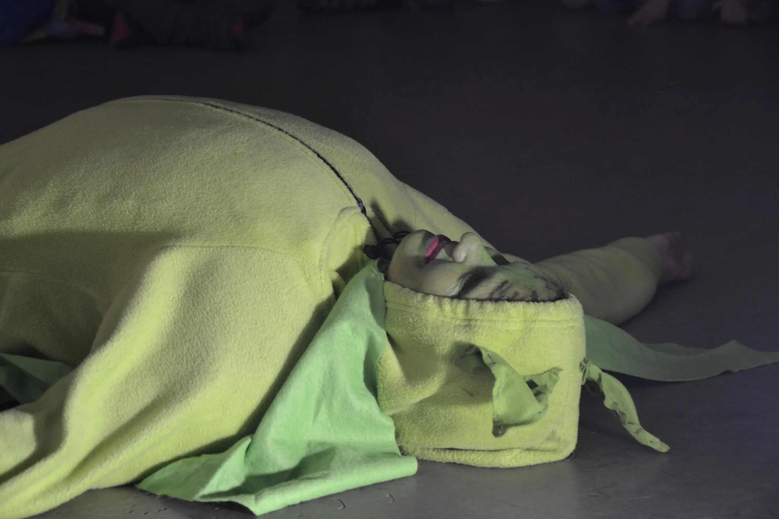 Un performer disteso a terra in un costume verde brillante, con un cappuccio che copre la testa, in un'atmosfera di danza o teatro.