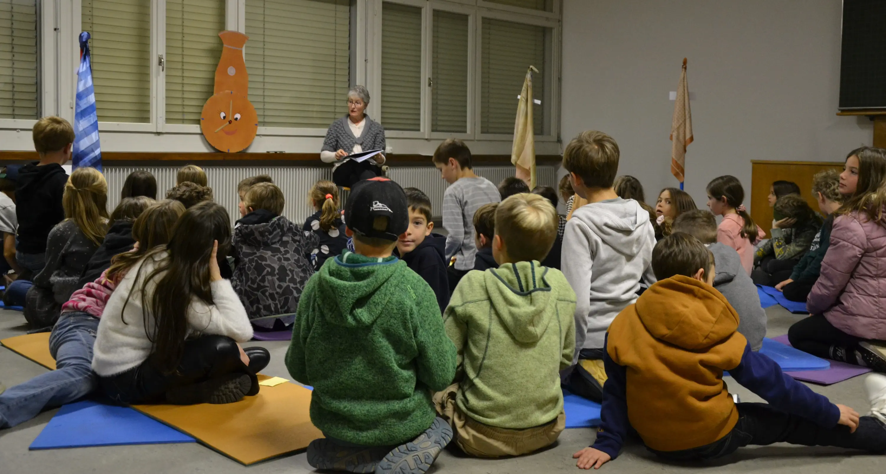 Un gruppo di bambini seduti su tappetini ascolta una donna che legge un libro in un ambiente interno. Sullo sfondo ci sono bandierine e decorazioni appese alle pareti.