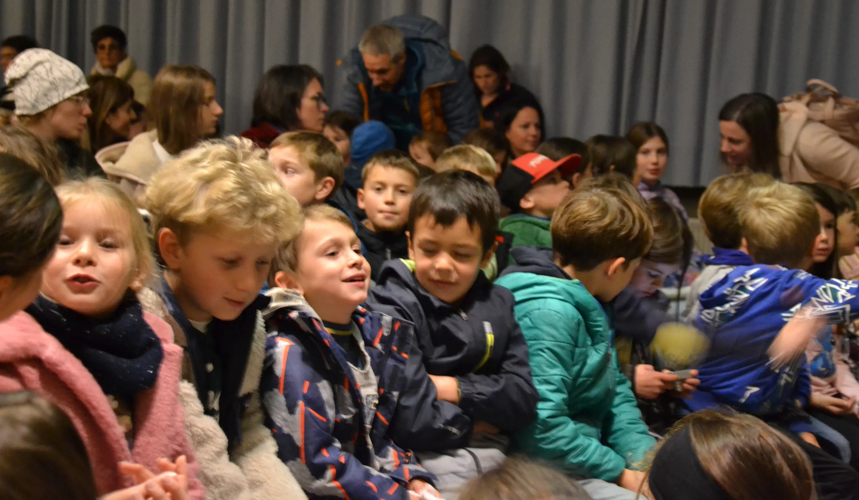 Un gruppo di bambini seduti in fila, sorridenti e impegnati in conversazioni, mentre adulti li osservano dal retro. La scena è vivace e informale.