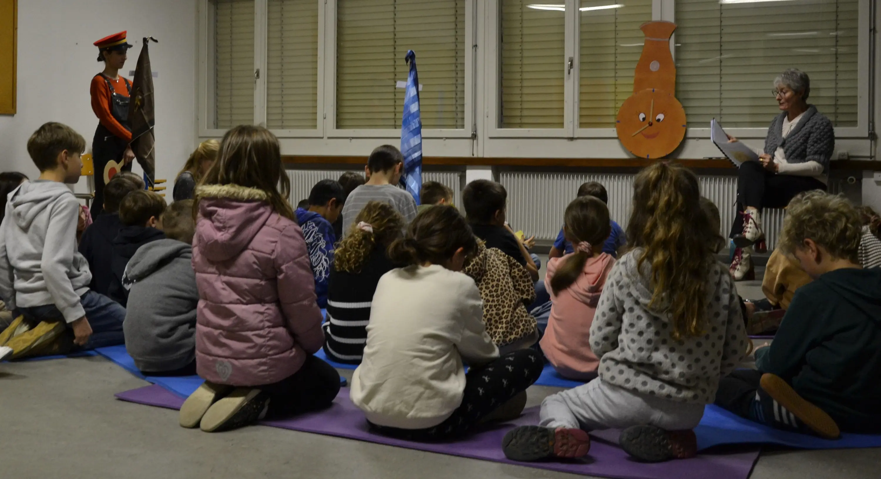 Un gruppo di bambini seduti su tappeti sul pavimento, ascolta una donna anziana che legge un libro. Un'altra persona, vestita in uniforme, sta in piedi accanto a una bandiera.
