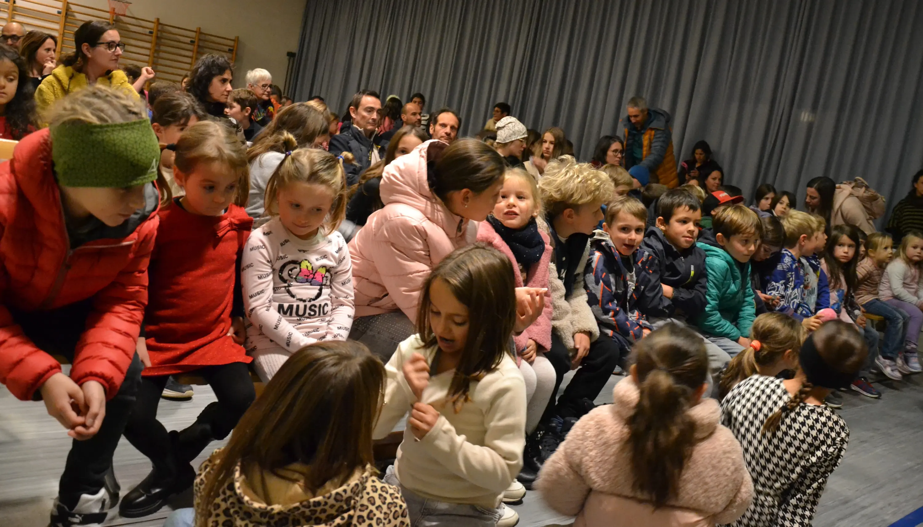 Un gruppo di bambini seduti in un'aula, alcuni indossano giacche colorate. Gli adulti si trovano sullo sfondo, mentre i bambini sono assorti in diverse attività. La scena sembra essere durante un evento o uno spettacolo.