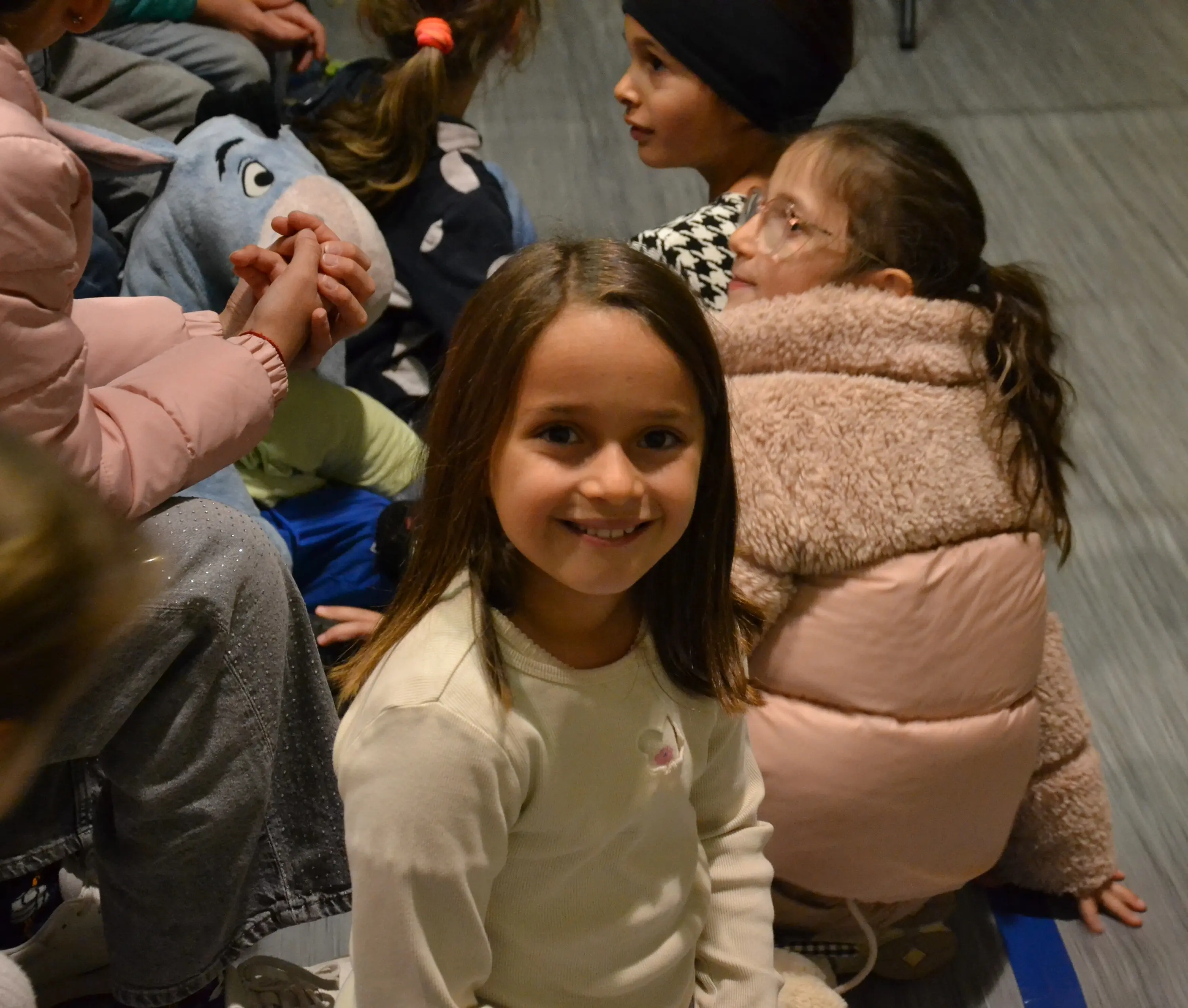 Una bambina sorridente seduta tra un gruppo di altri bambini, indossa una maglietta chiara e ha i capelli lunghi e castani. Sullo sfondo si vedono altri volti, alcuni dei quali girati.