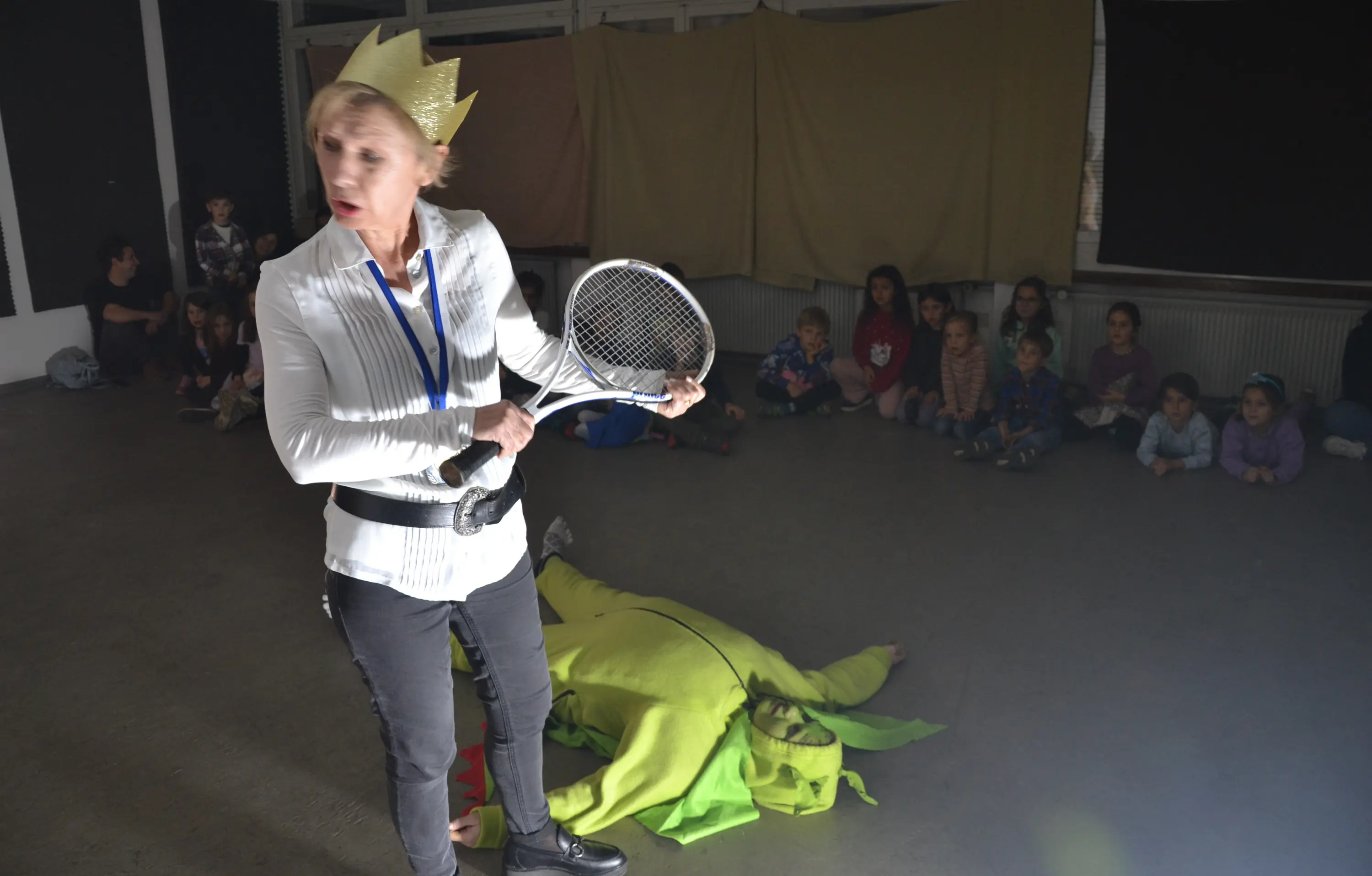 Una donna con una corona d'oro e una racchetta da tennis si volta in modo energico, mentre un uomo vestito da creatura verde è sdraiato a terra. Sullo sfondo, un gruppo di bambini osserva attentamente la scena.