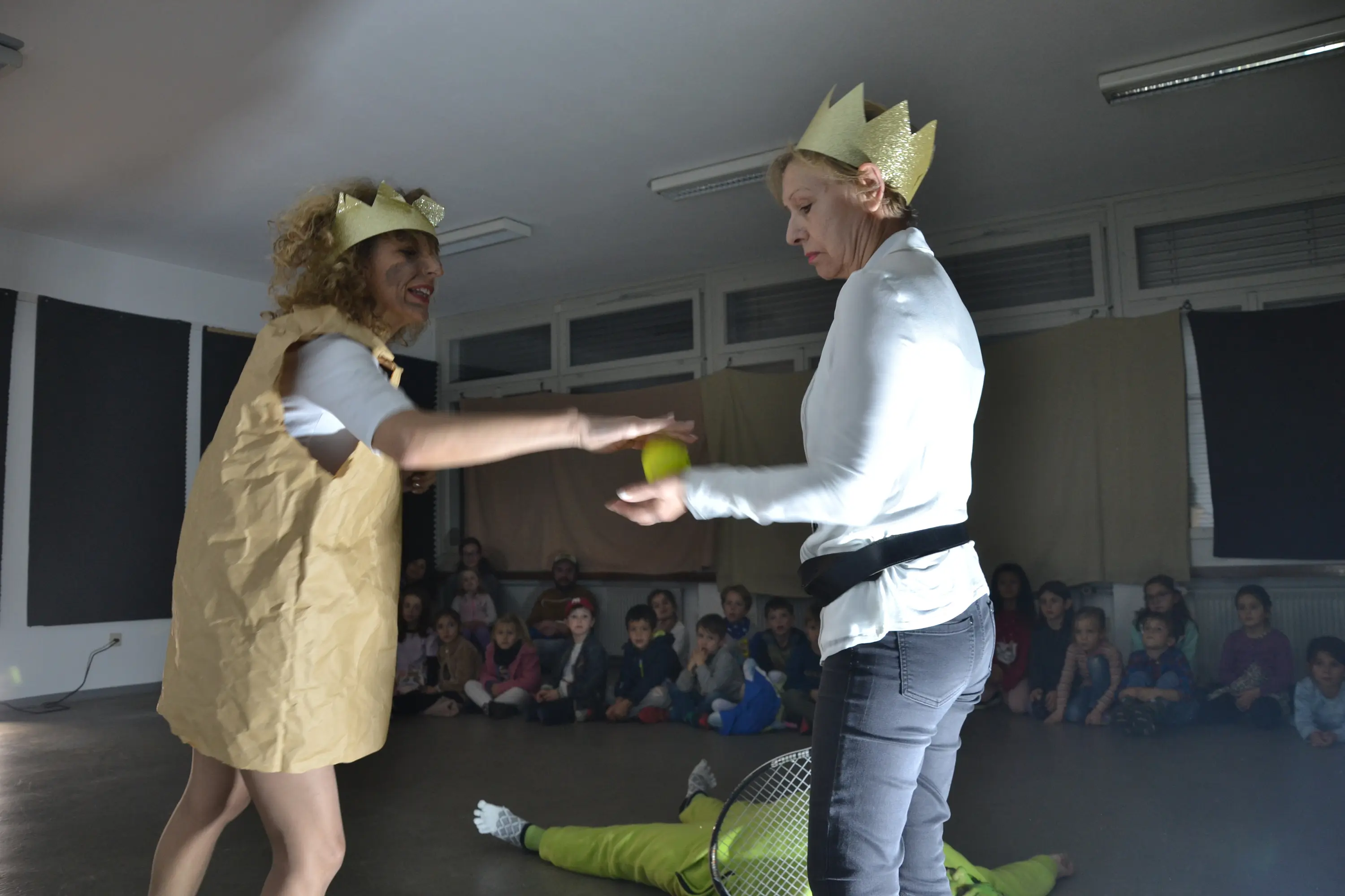 Due donne in costume, una con una corona di carta, si scambiano una palla gialla. Sullo sfondo, un gruppo di bambini osserva attentamente.