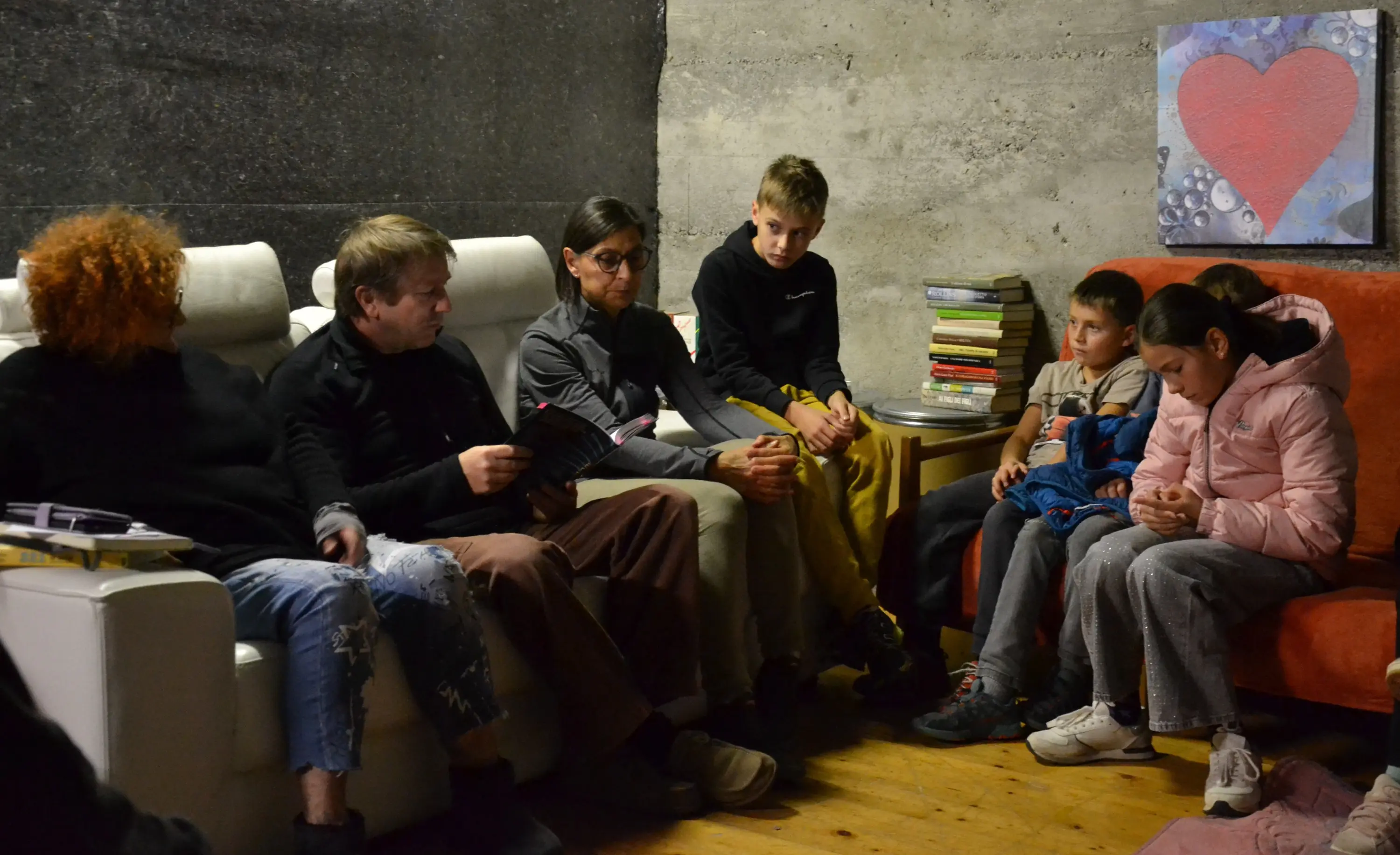 Un gruppo di persone sedute in una stanza, alcune mentre ascoltano un uomo che legge. Due bambini sono seduti in primo piano, concentrati, mentre gli adulti, vestiti in modo casual, sono disposti attorno a loro su divani. Sullo sfondo, ci