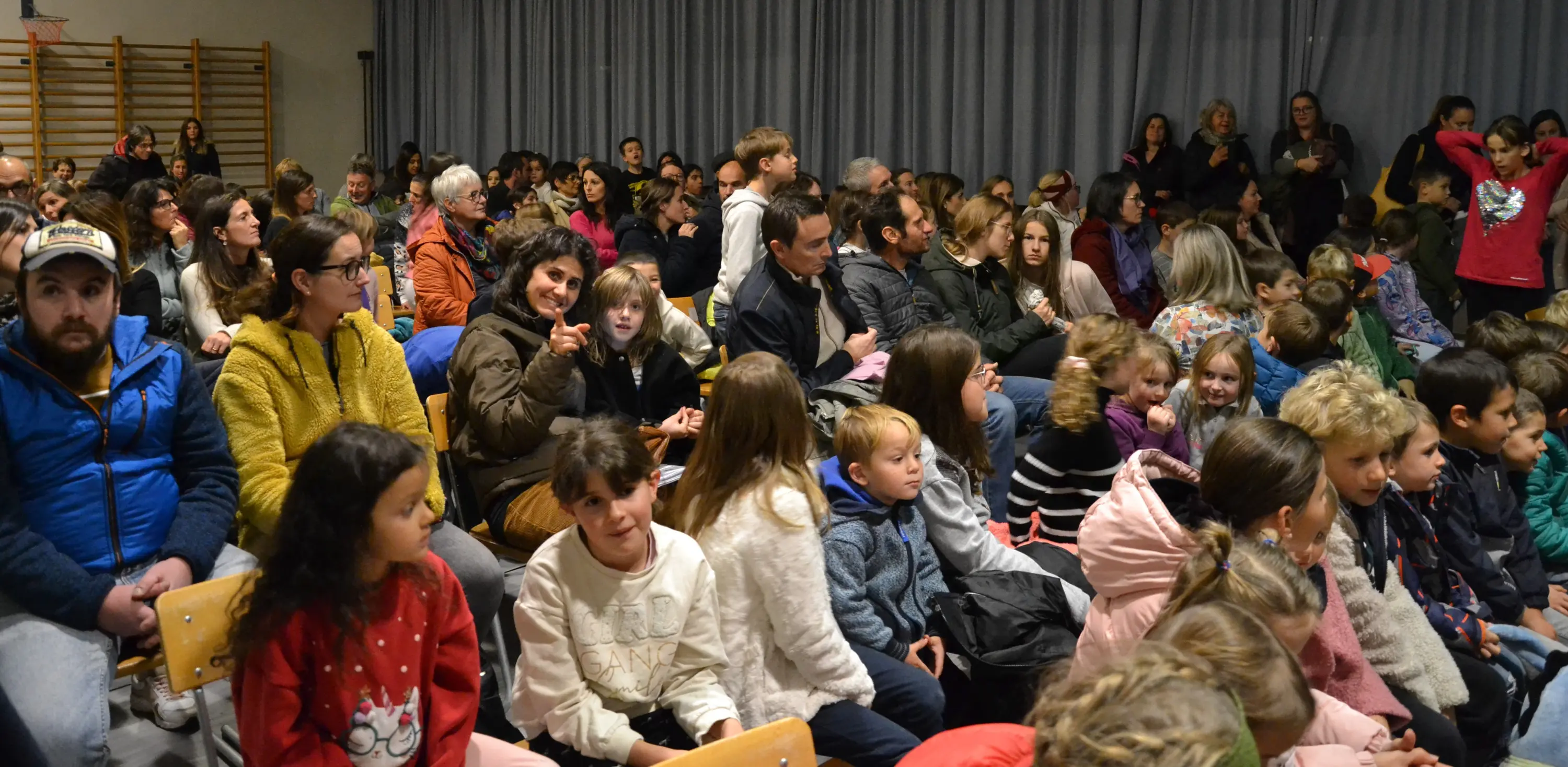 Un pubblico affollato, composto da adulti e bambini, seduto in una sala per un evento. Molti sorridono e chiacchierano, con un'atmosfera vivace e conviviale.