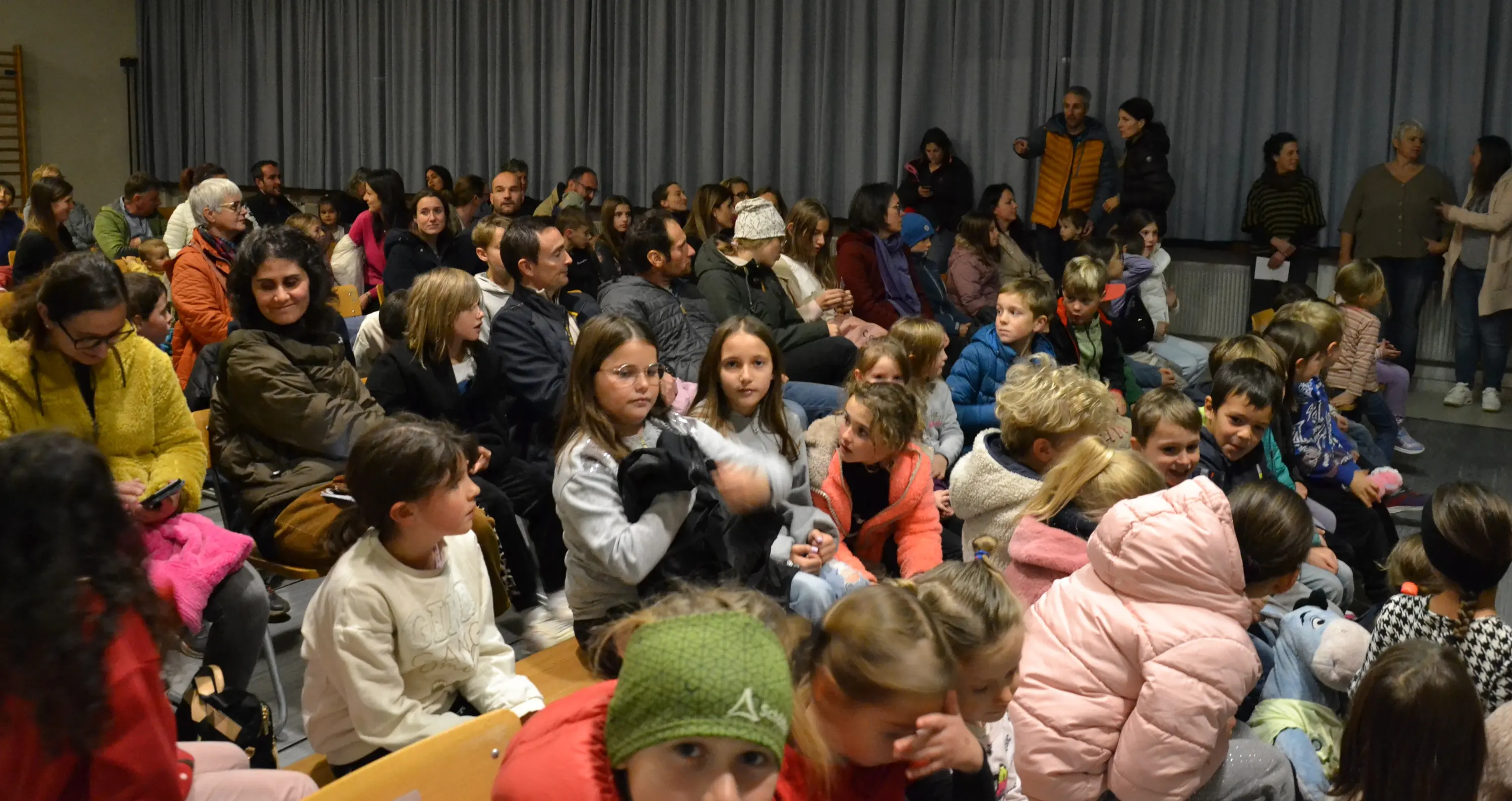 Una folla di adulti e bambini seduti in una sala, alcuni guardano verso la telecamera. Molti indossano giacche colorate e prendono parte a un evento o a una riunione.